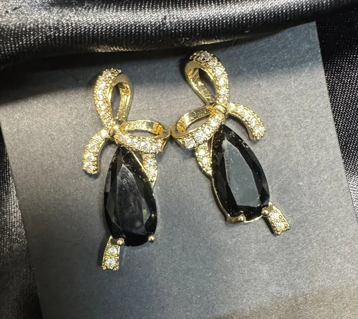 Gold bow-shaped earrings with black teardrop stones on a dark fabric background