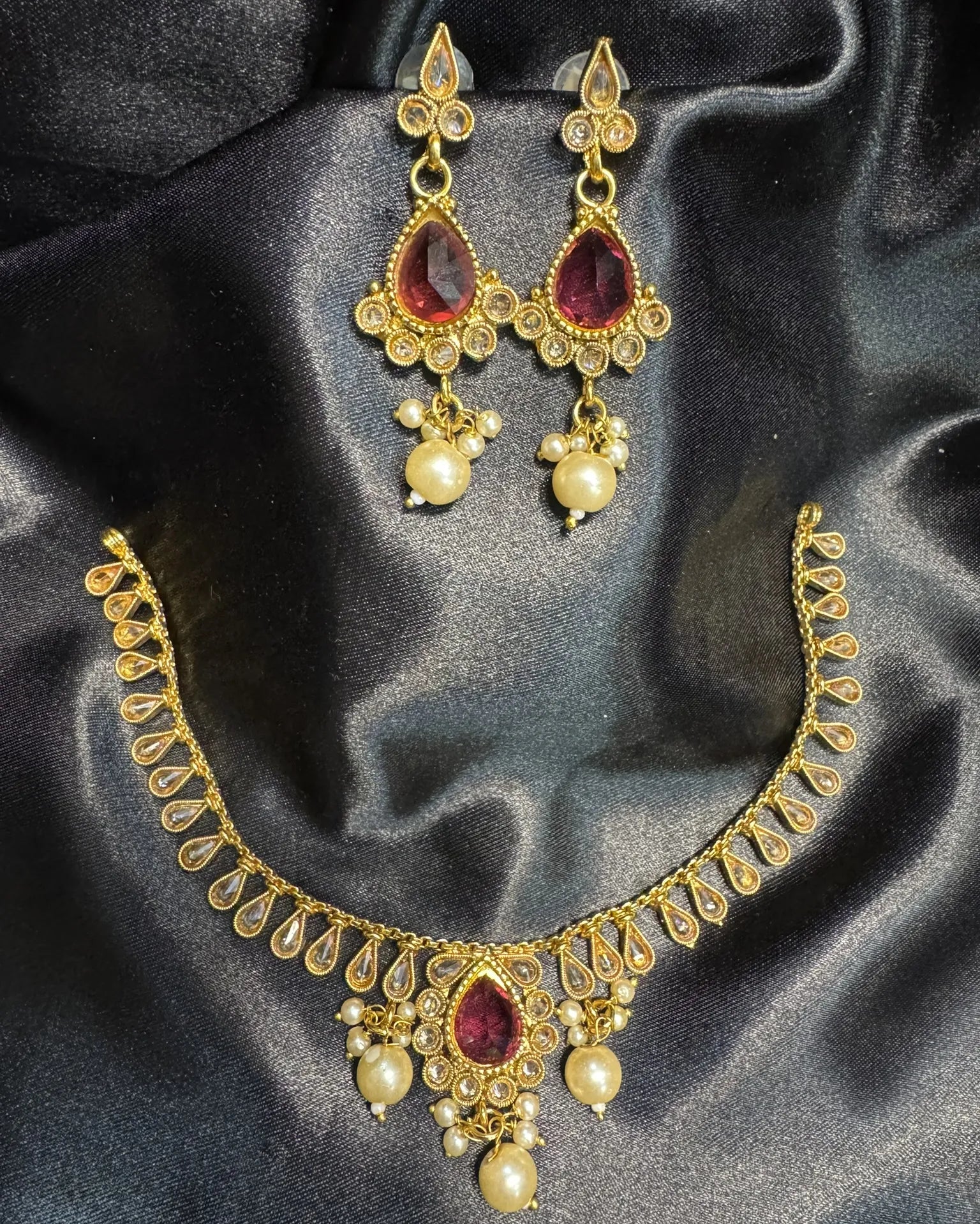 Gold necklace and earrings with hot pink gemstones and pearl-like beads on a dark satin background