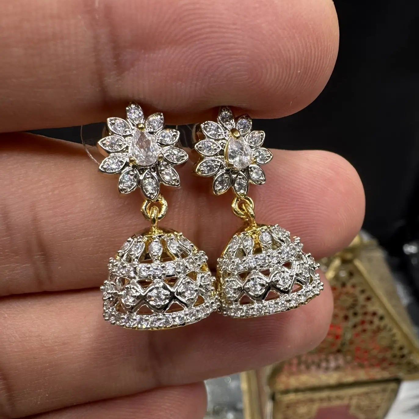 Pair of Pakistani style jhumka earrings with clear gemstones and a gold base held between fingers with a blurred background 