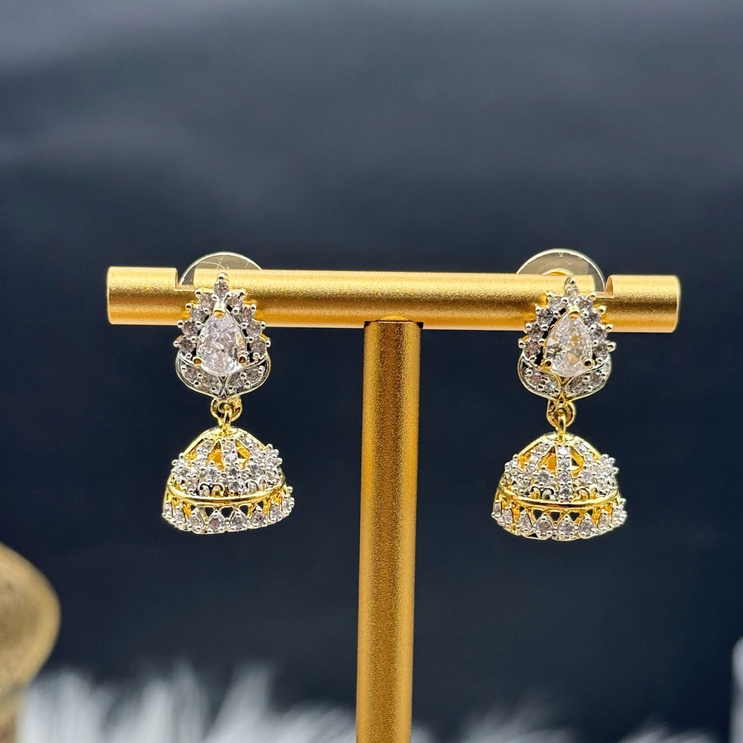 Pair of Pakistani style jhumka earrings with clear gemstones and a gold base on a gold stand against a dark background