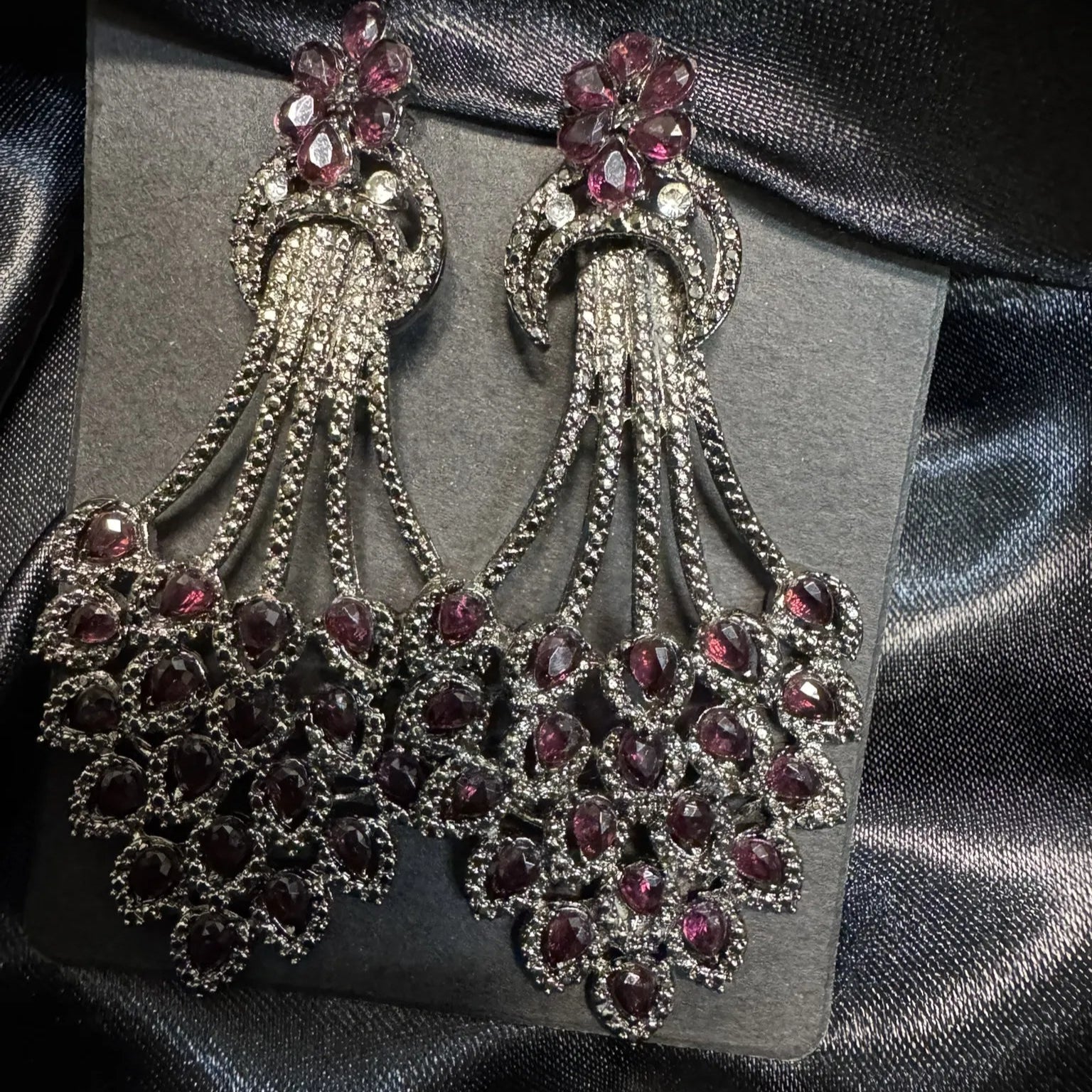 Pair of ornate earrings with hotpink gemstones on a gray card against a black satin background
