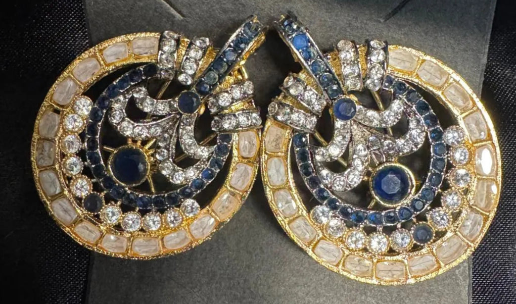 Pair of ornate earrings with blue gemstones on a gray card with a dark fabric background
