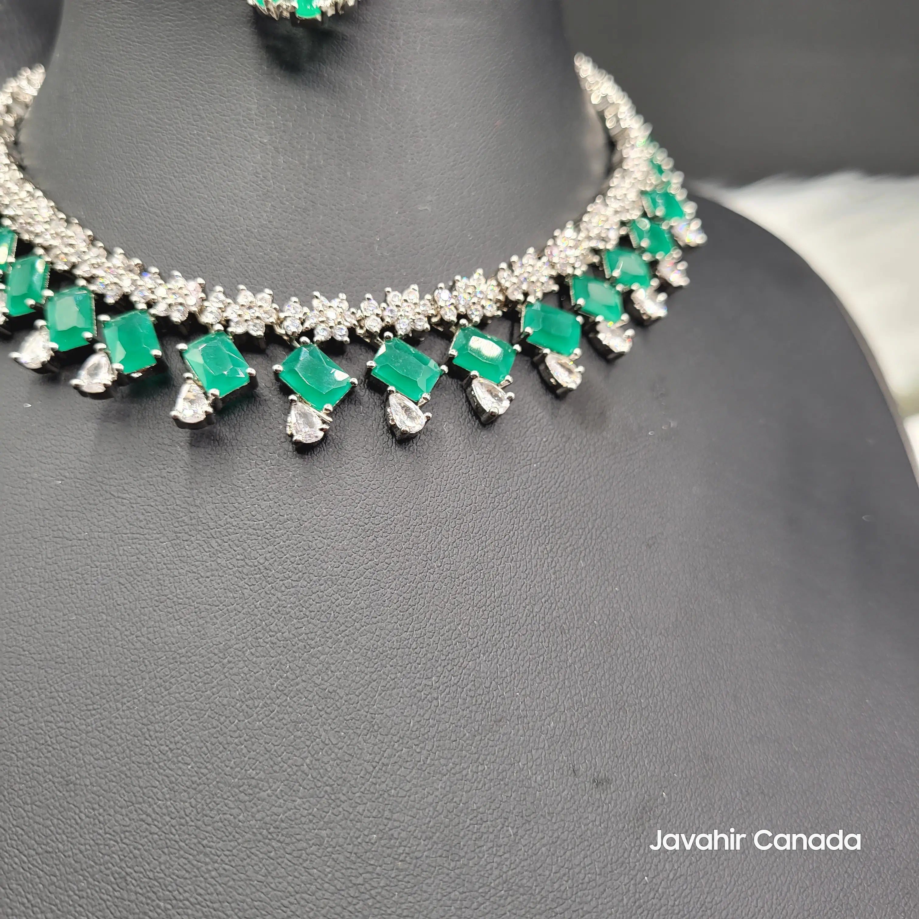Close-up of JV19 emerald necklace showcasing green rectangular stones with teardrop CZ accents on a black display.