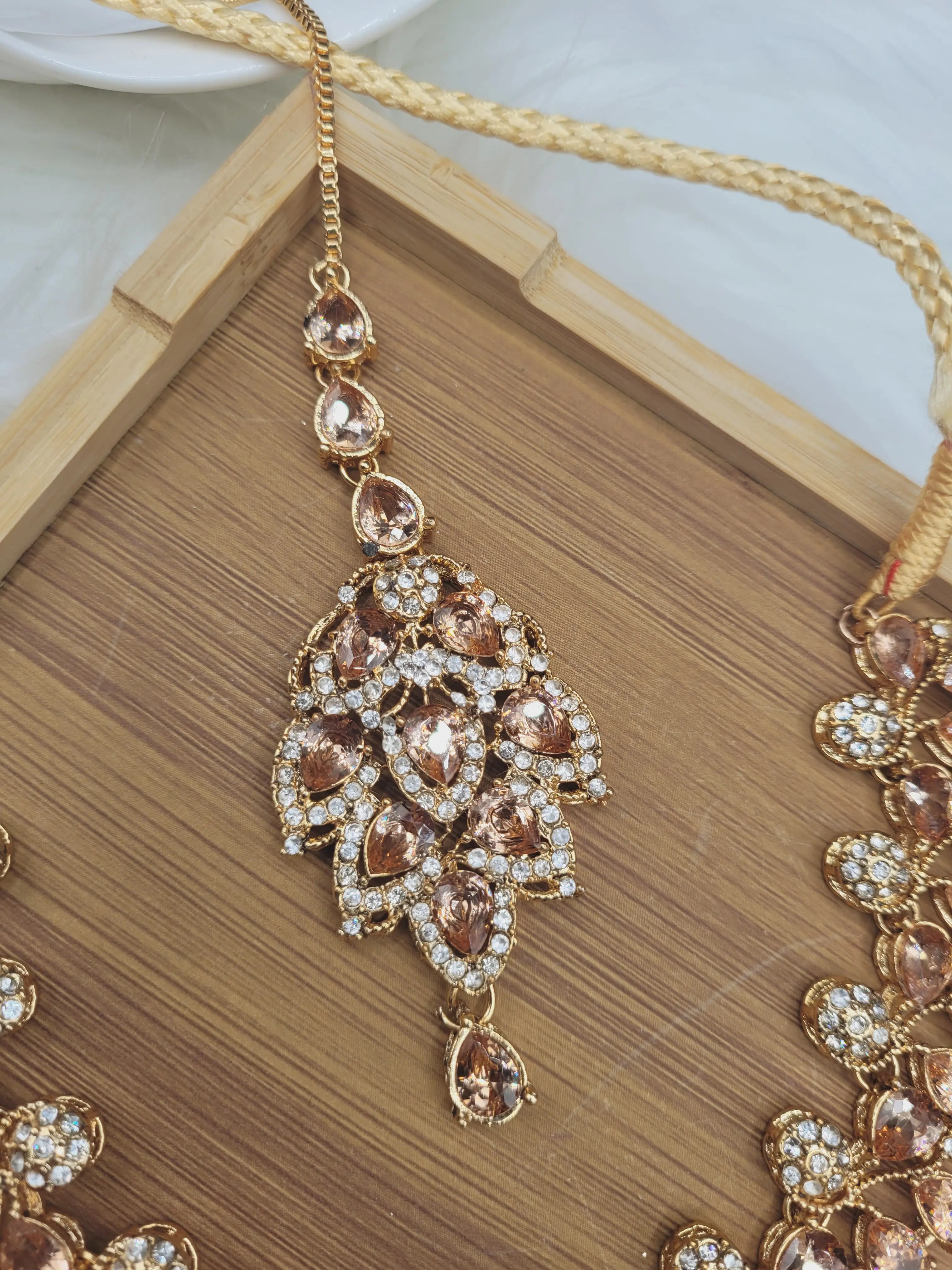 Champagne variant maang tikka from JV20 set, featuring pear-cut stones and round CZ detailing, laid flat on wooden tray.