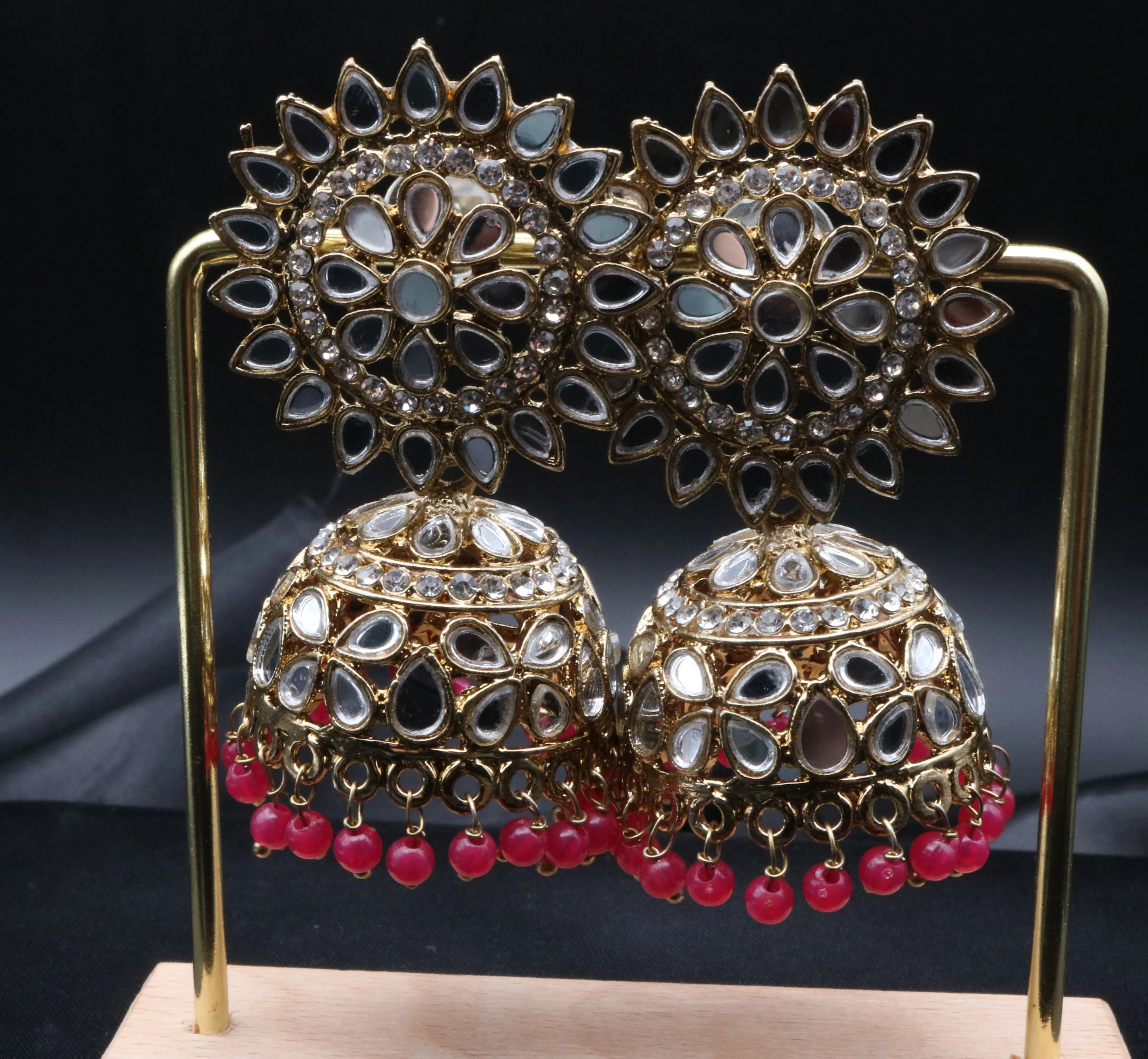 Pair of gold plated with magenta beads kundan jhumka earrings on a stand against a gray background