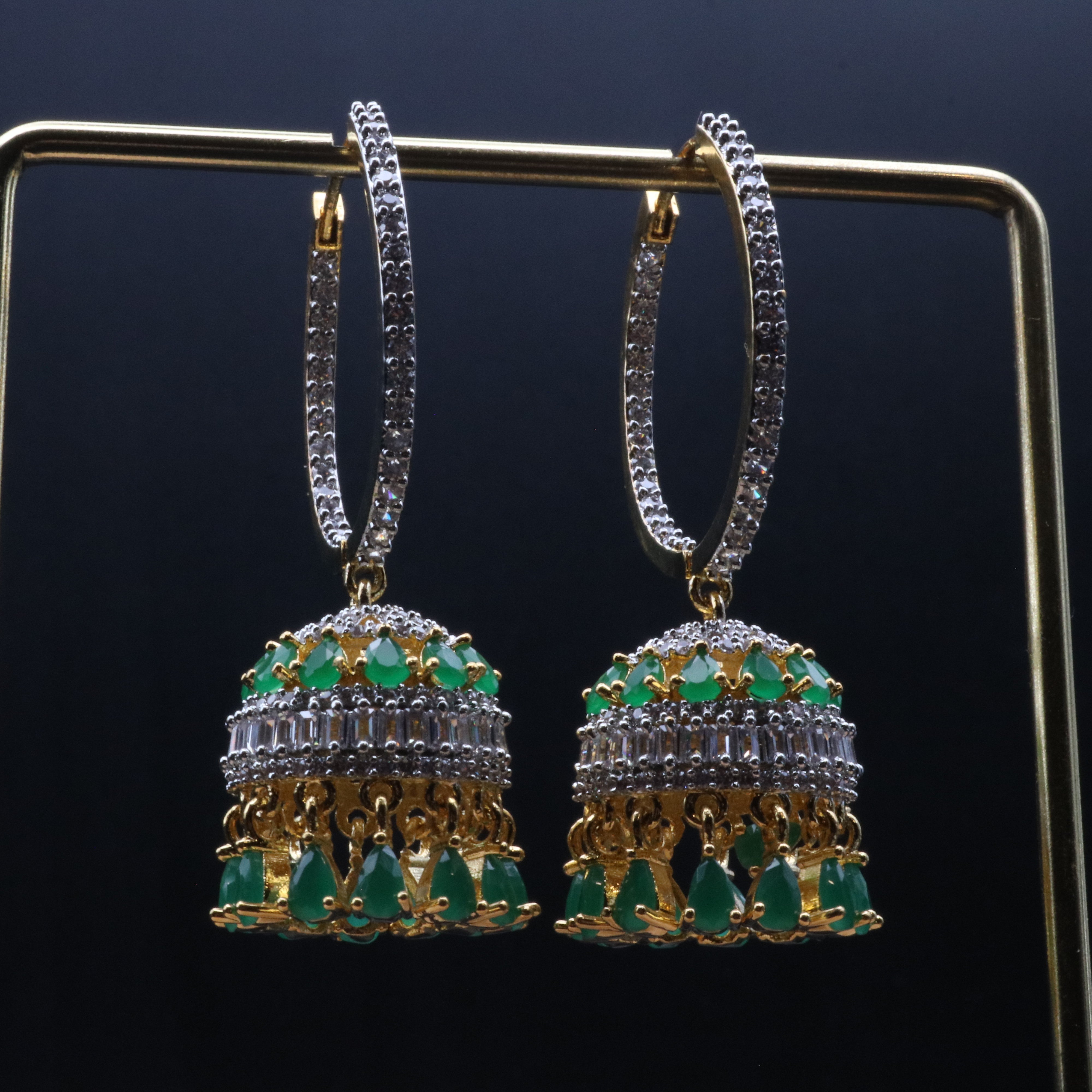 A pair of gold-colored jhumka earrings featuring green and clear gemstones, displayed against a dark background.