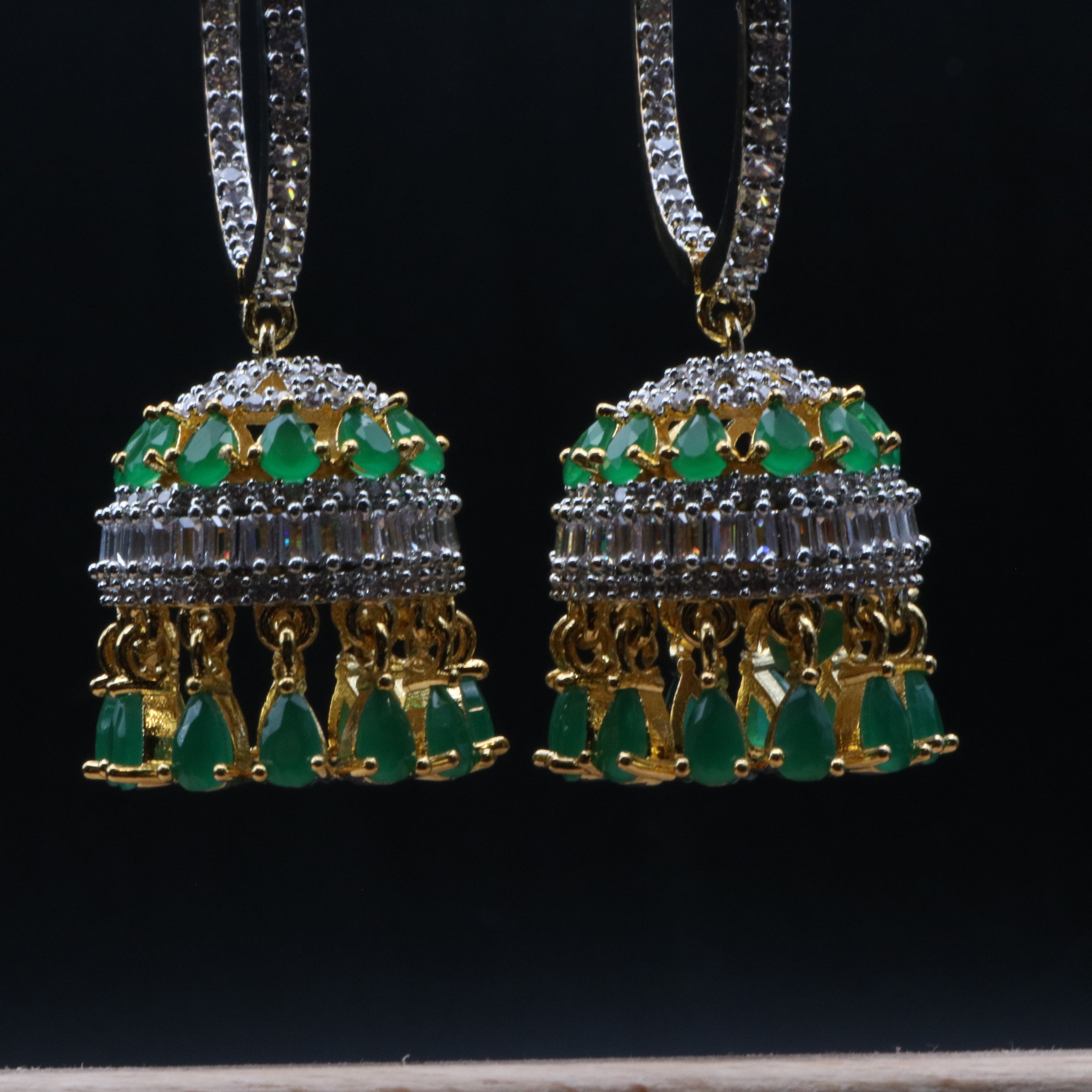 close view of jhumkay for a pair of gold-colored jhumka earrings featuring green and clear gemstones, displayed against a dark background.