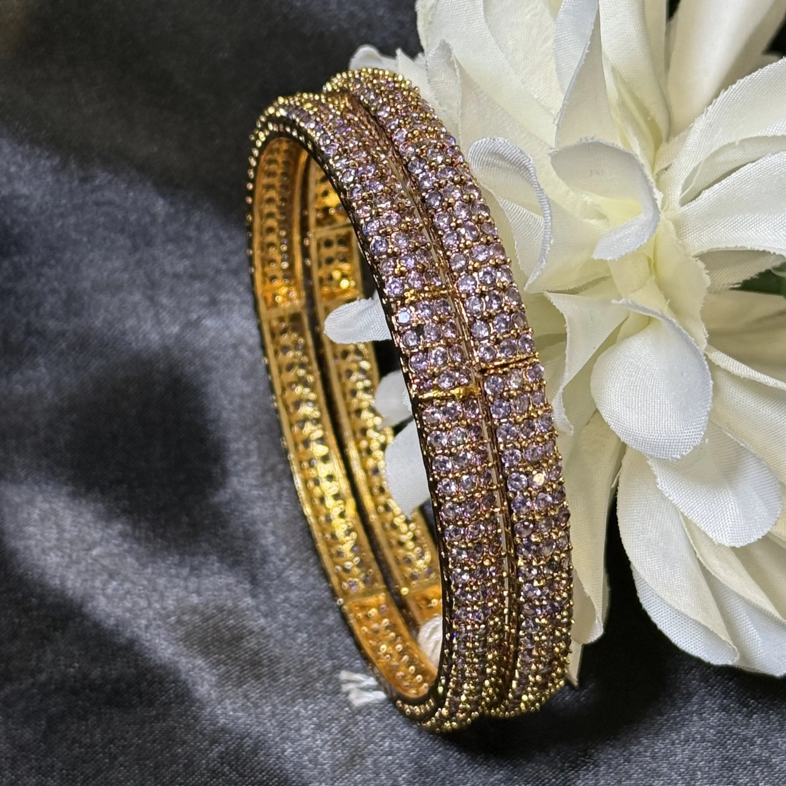Gold bangles with gemstones on a white flower against a gray background