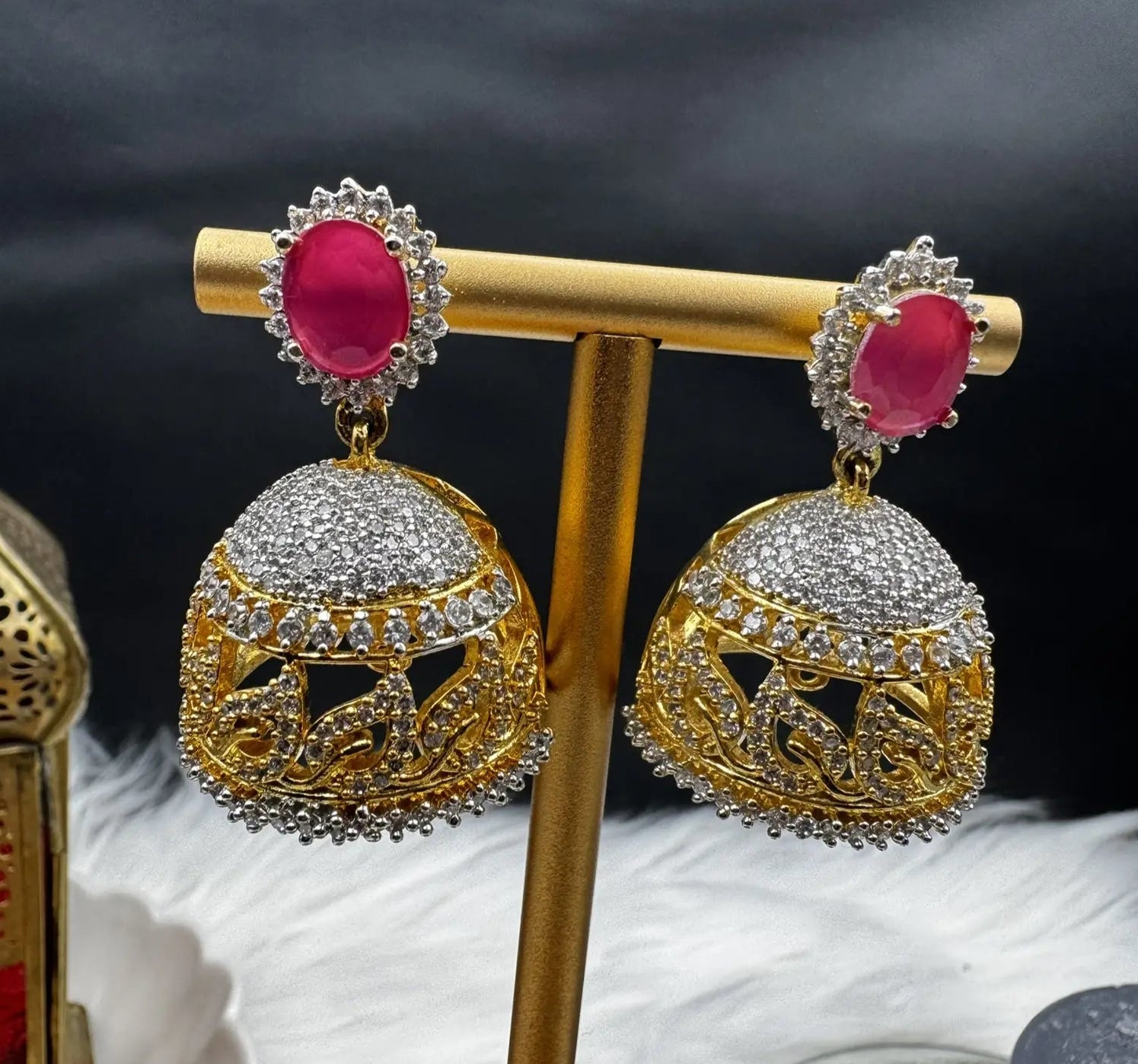 another anlge of goldplated and nano zarcon Pakistani style jhumka earrings with hotpink gemstones on a gold stand against a dark background. Jarao based design 
