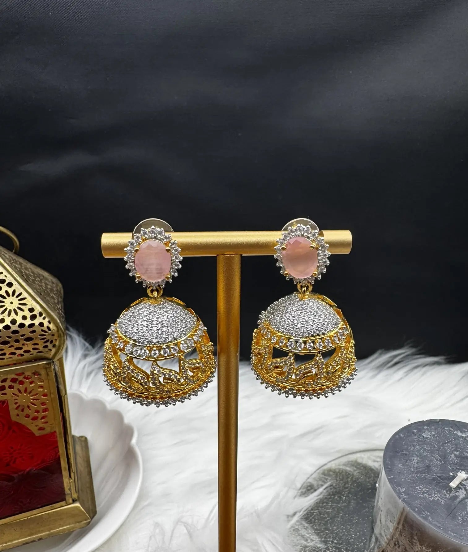 slight tilted front angle of goldplated and nano zarcon Pakistani style jhumka earrings with pink gemstones on a gold stand against a dark background. Jarao based design 