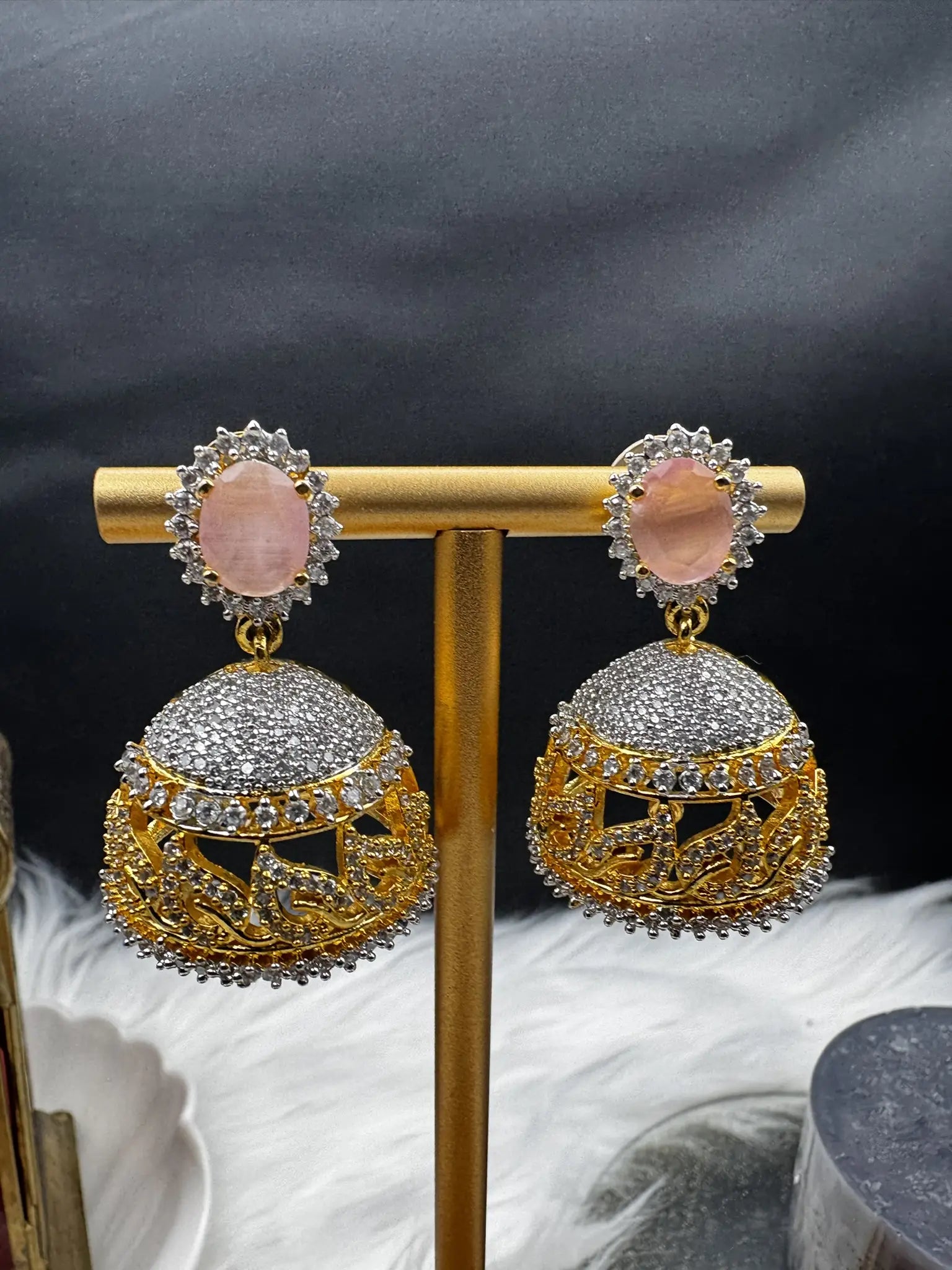 closer angle of goldplated and nano zarcon Pakistani style jhumka earrings with pink gemstones on a gold stand against a dark background. Jarao based design 