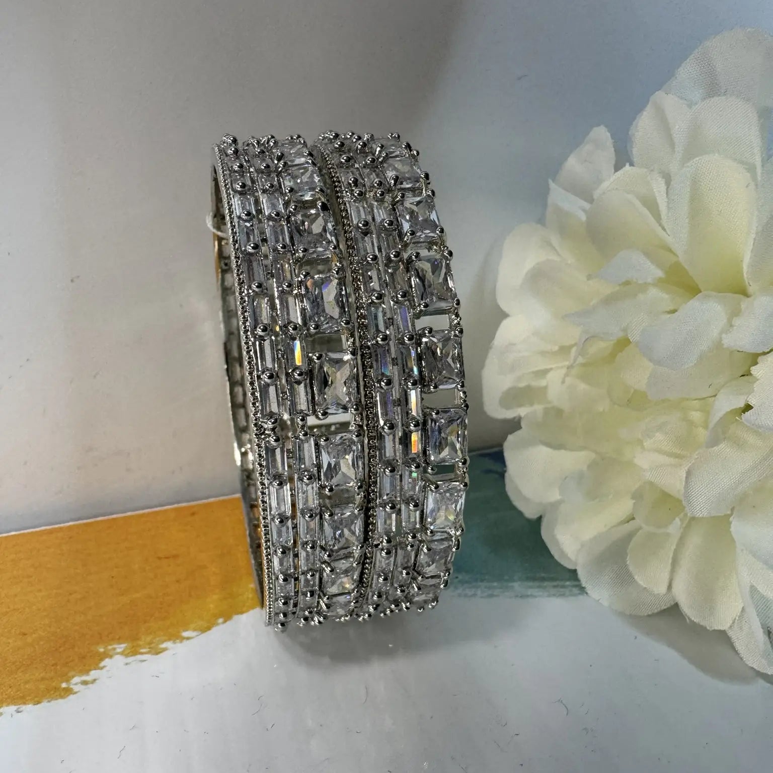stainless steel bangles adorned with shimmering American diamonds and clear gemstones on a white surface with a yellow object and white flower.