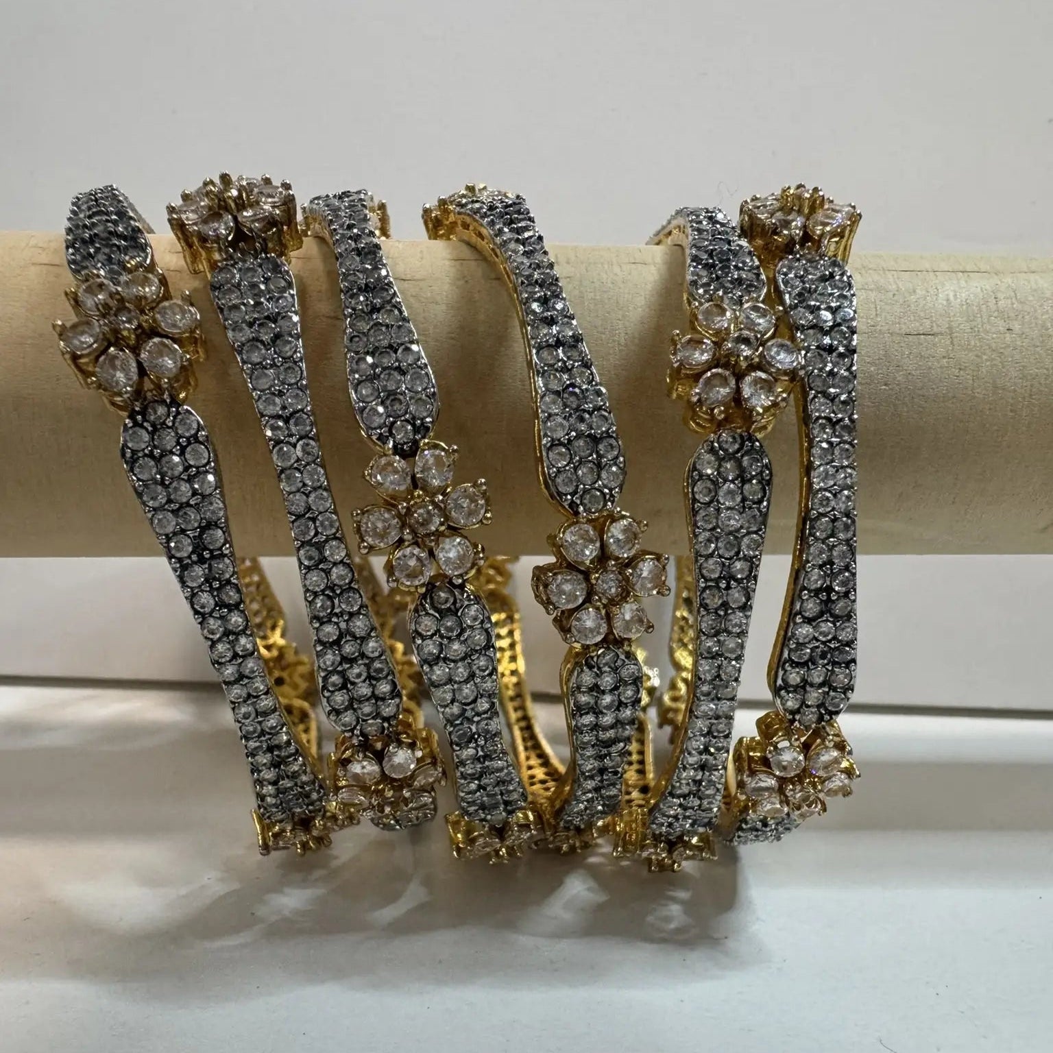 Set of gold-plated, American diamond bangles with flower patterns and clear gemstones on a wooden stick and white background