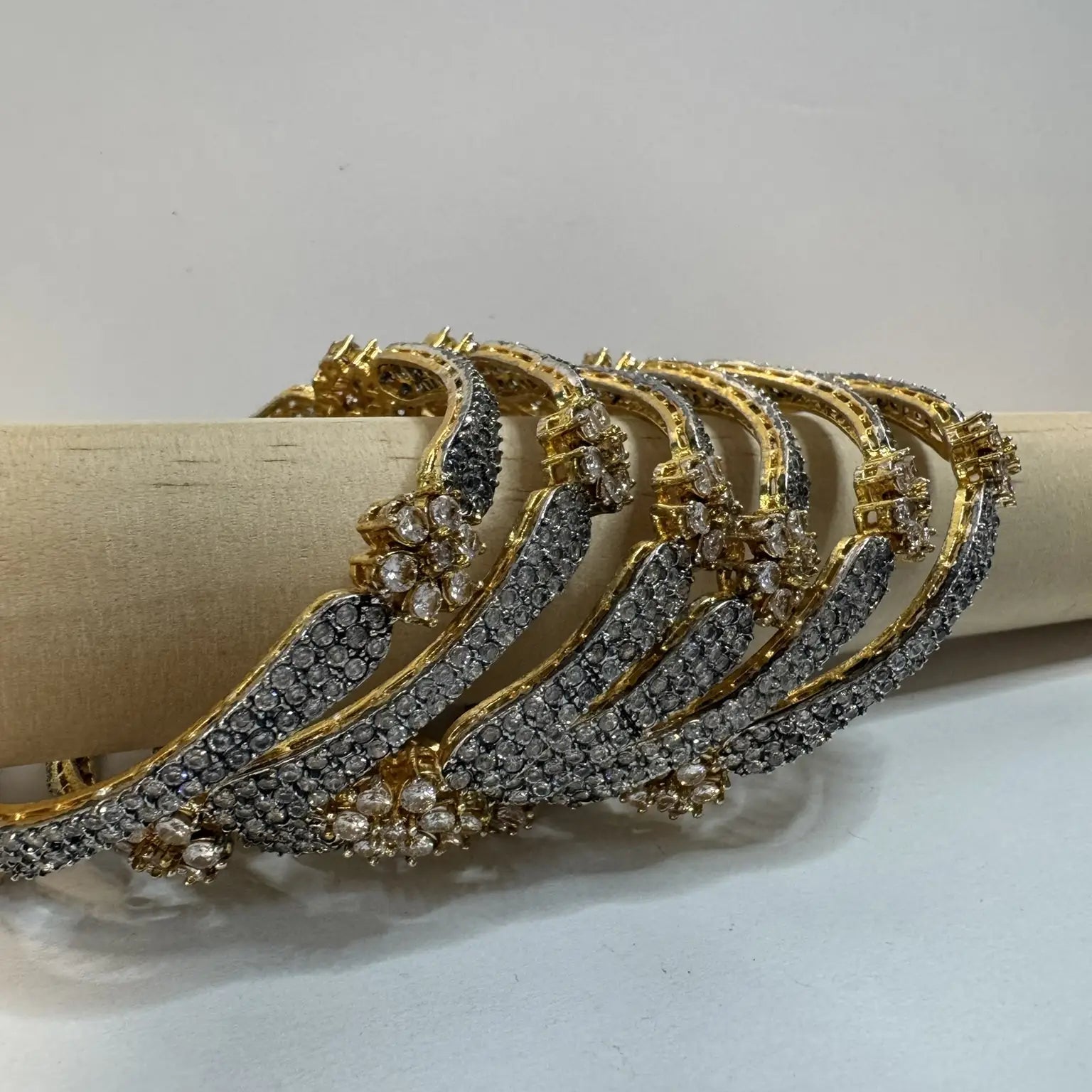 Gold-plated, American diamond bangles with flower patterns and clear gemstones on a white background and wooden stick