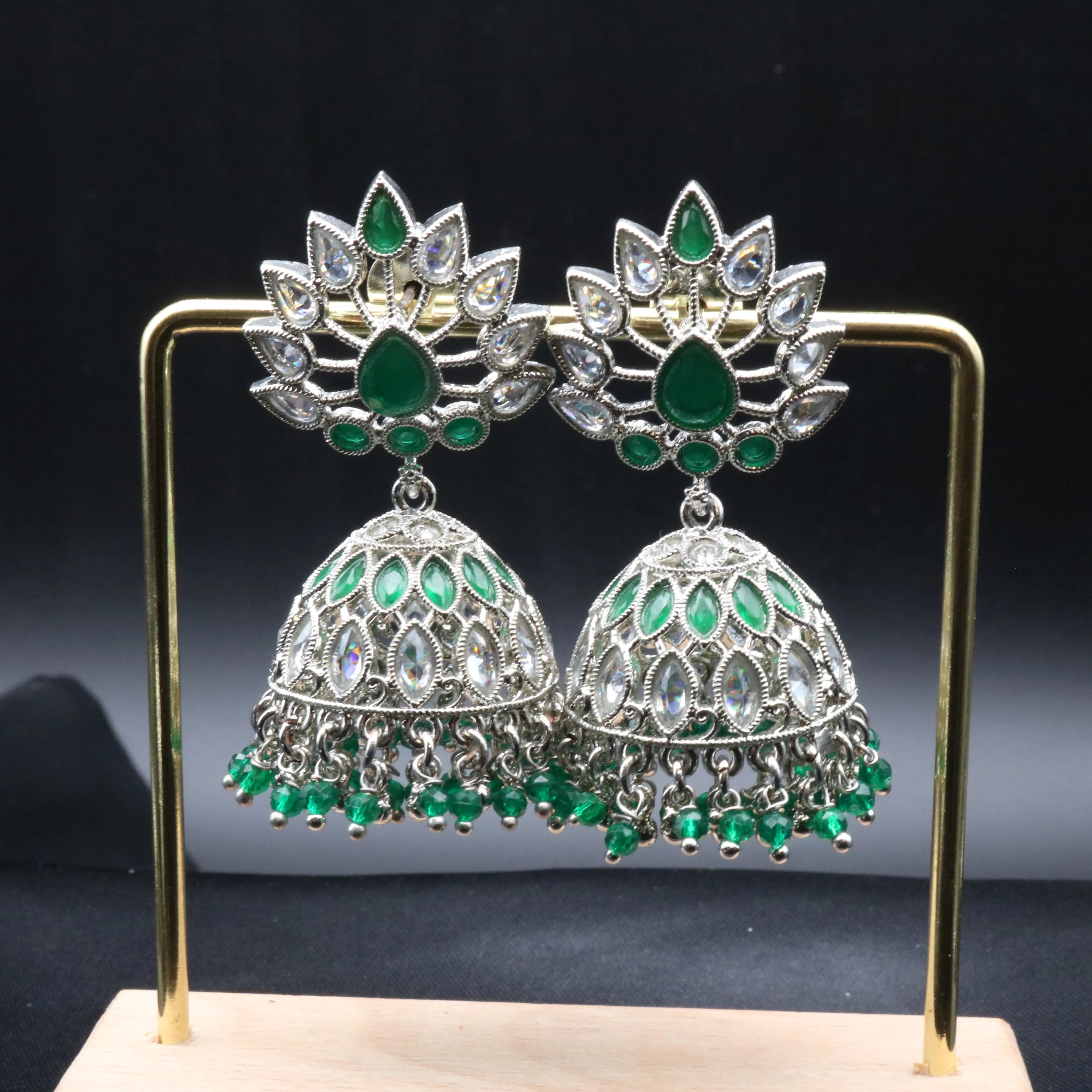 Pakistani style jhumka earrings with gsilver base and green and clear gemstones on a stand against a dark background