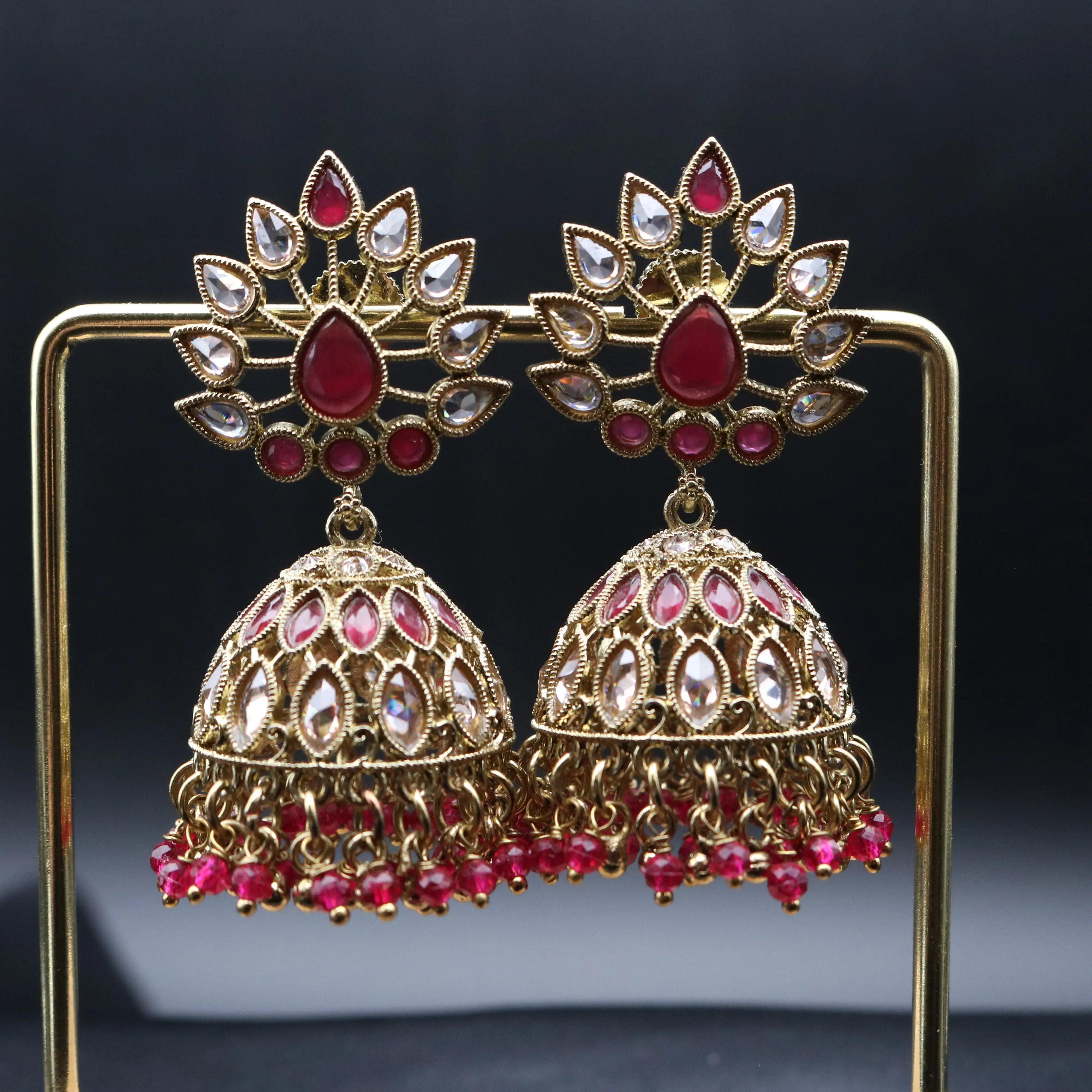 Pakistani style jhumka earrings with gold base and red and clear gemstones on a stand against a dark background