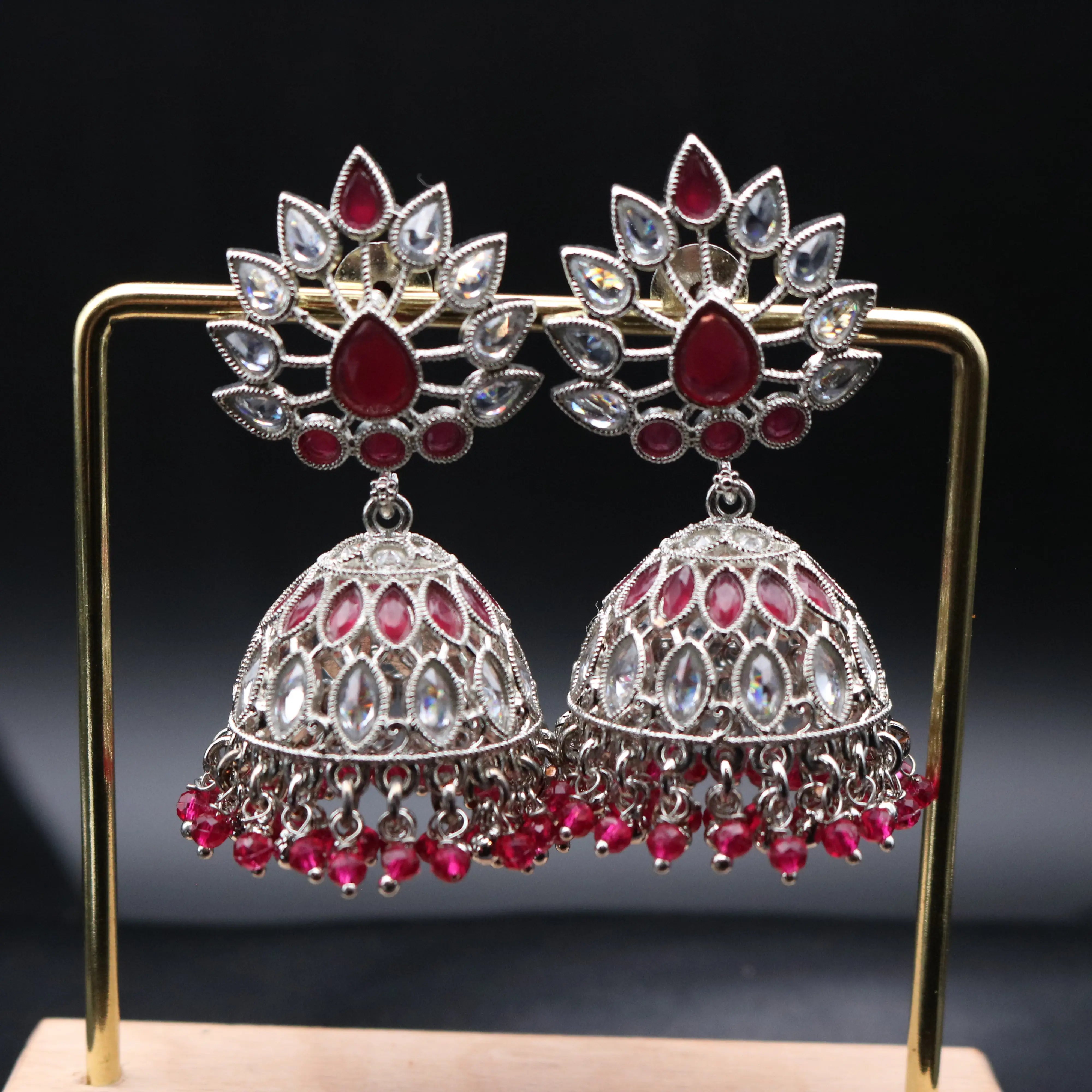 Pakistani style jhumka earrings with silver base and red and clear gemstones on a stand against a dark background