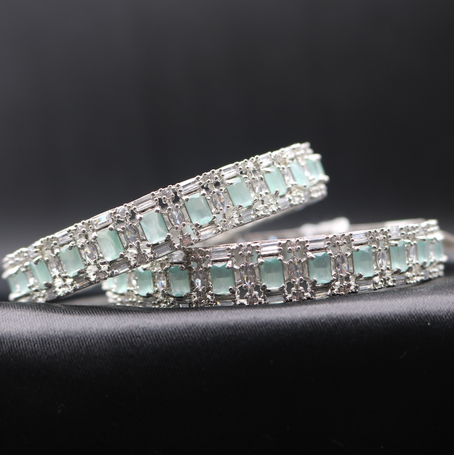 This image displays a pair of elegant castle-themed bangles. Each bangle is adorned with sparkling teal gemstones set in a meticulously crafted metal frame, showcasing a pattern that resembles a castle wall.