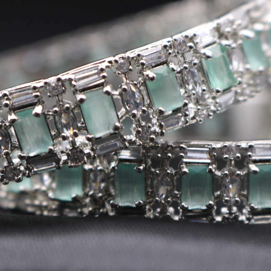 Closer view of a pair of elegant castle-themed bangles. Each bangle is adorned with sparkling teal gemstones set in a meticulously crafted metal frame, showcasing a pattern that resembles a castle wall.
