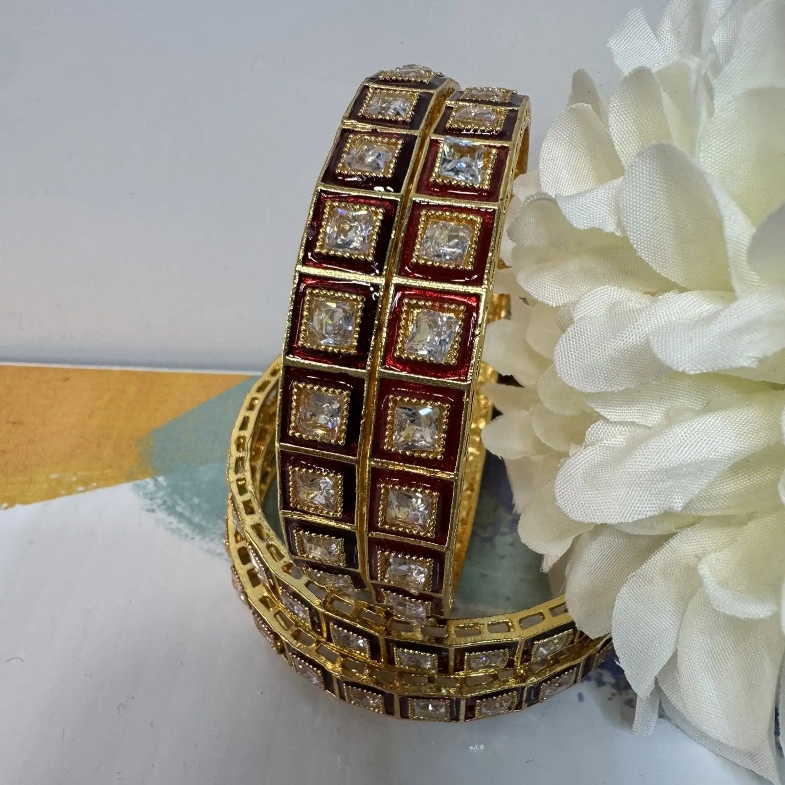 Red and gold, gold-plated bangles with square gemstones next to a white flower on a light surface