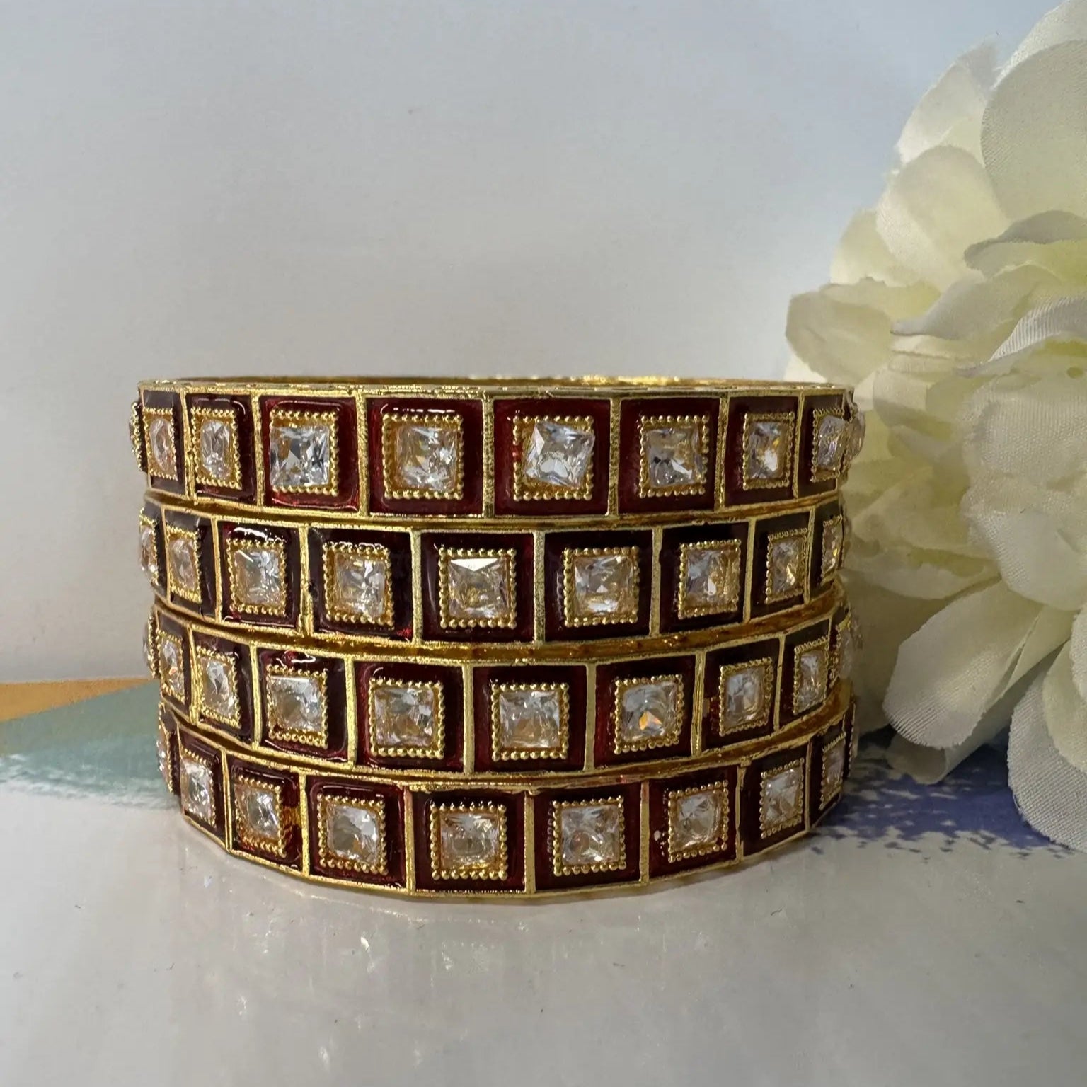 Red and gold, gold-plated bangles with square gemstones on a white surface with a flower.