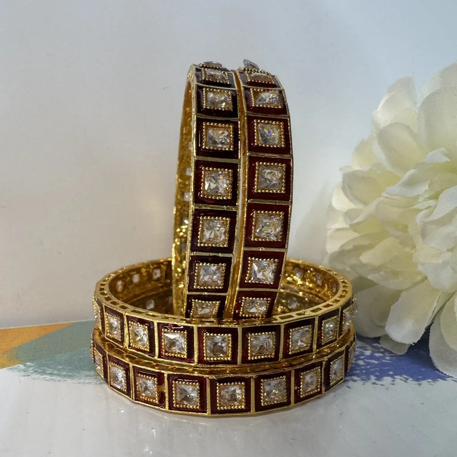 Red and gold, gold-plated bangles with square gemstones on a white surface with a flower in the background