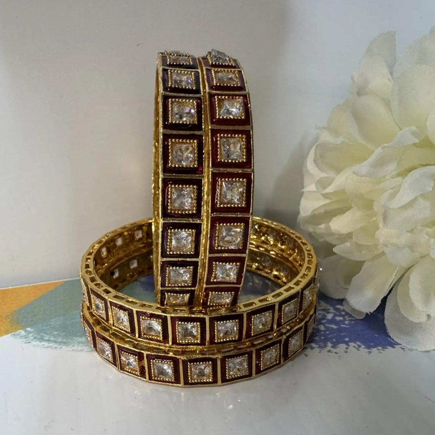 Red and gold, gold-plated bangles with square gemstones on a white surface with a white flower in the background