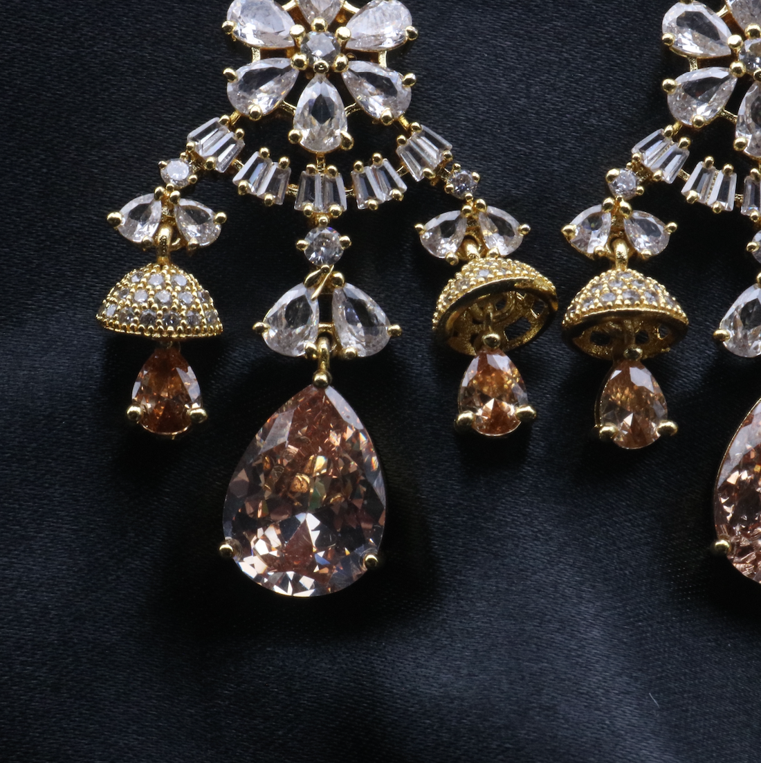 bottom view pair of stainless steel and gold-plated statement earrings with intricate designs and multiple layers of sparkling stones displayed against a dark background.
