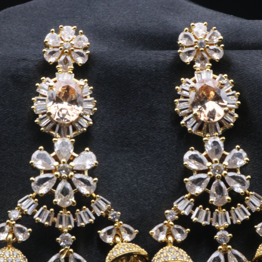 top view of pair of stainless steel and gold-plated statement earrings with intricate designs and multiple layers of sparkling stones displayed against a dark background.