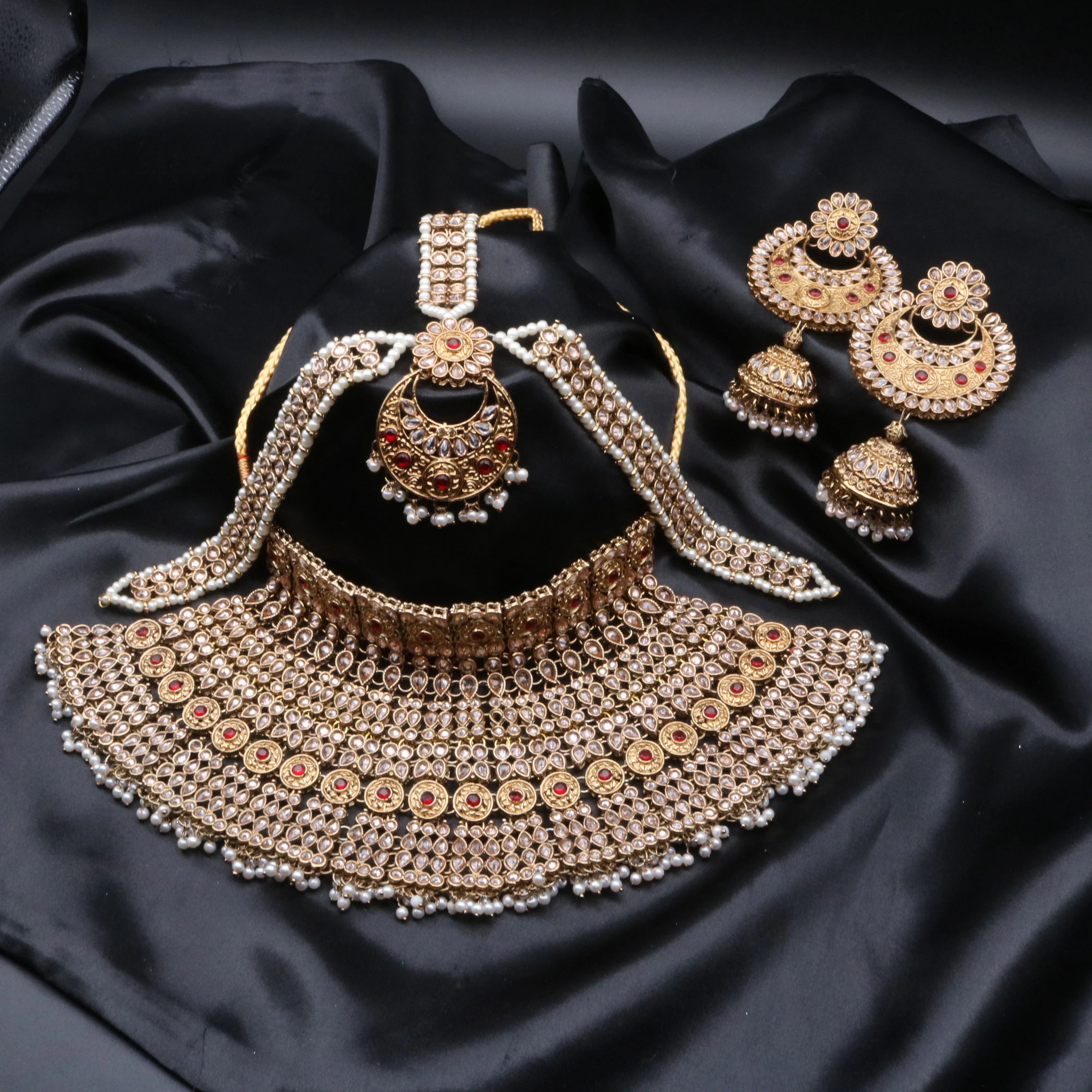 A gold-colored Pakistani style bridal set with intricate designs and pearl-like beads, displayed alongside a pair of matching jhumka earrings and matha patti on a dark fabric background.