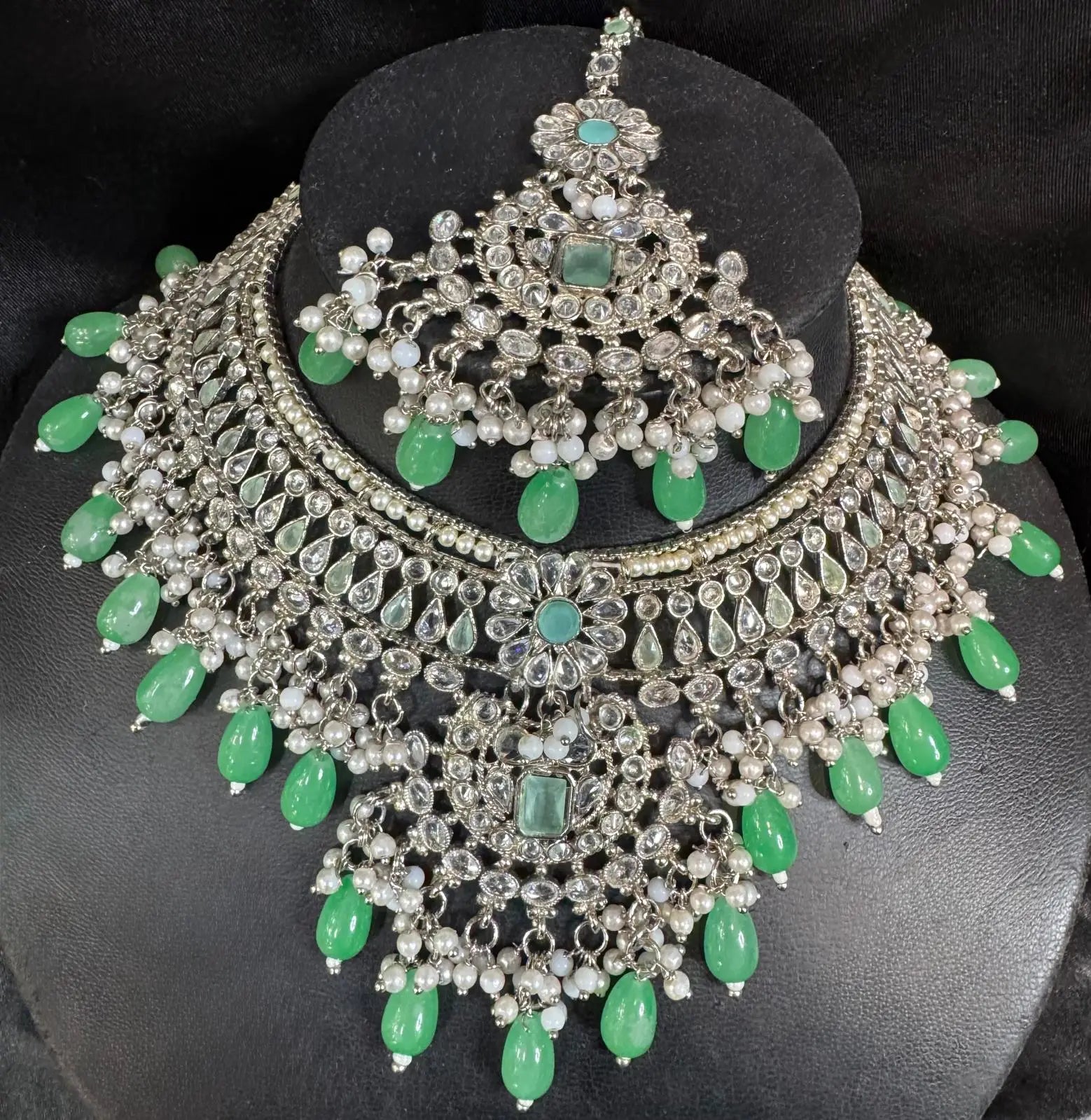 JV157 necklace set displayed on stand: heavy choker with kundan stones, green glass drops, pearl detailing, and matching maang tikka.