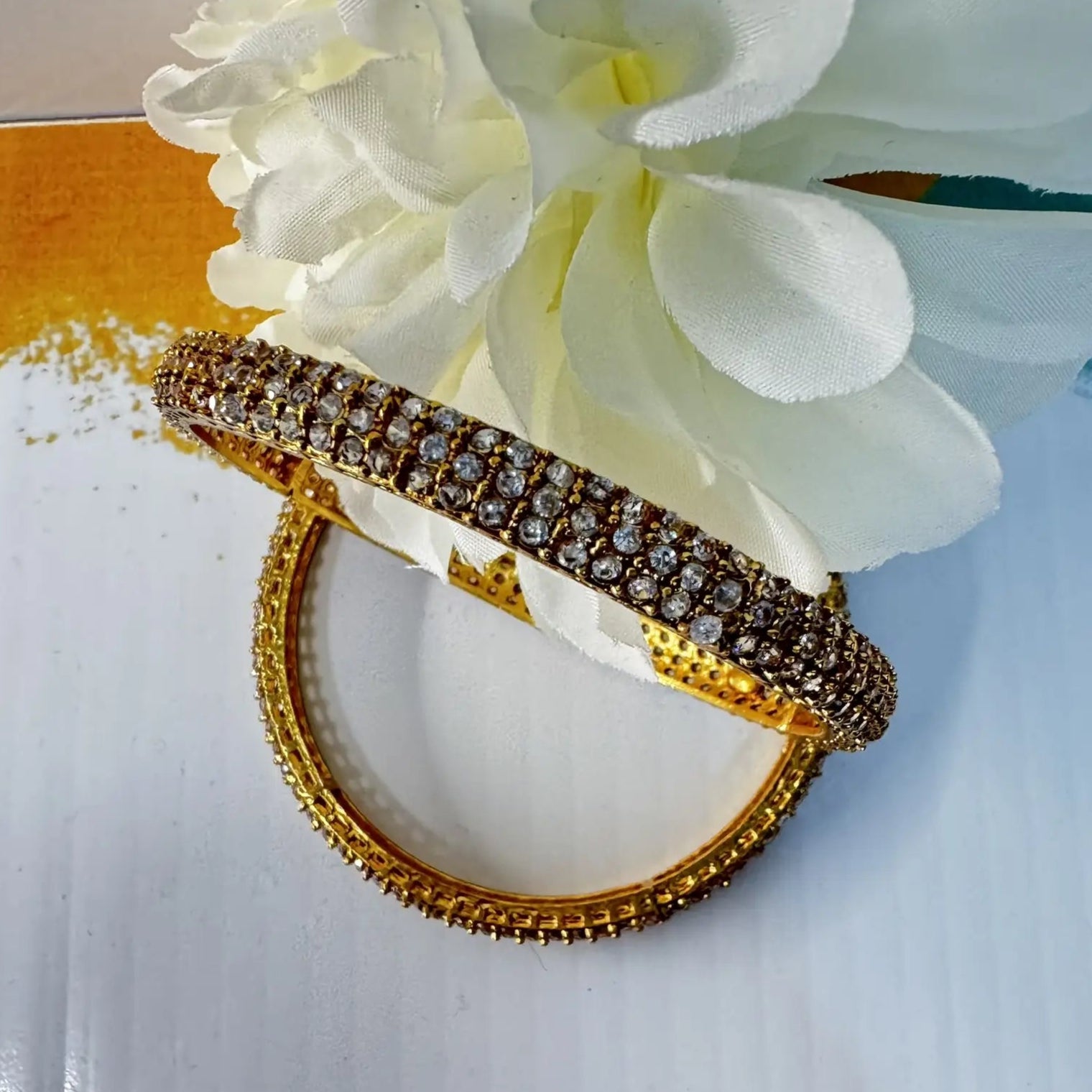 Gold plated bangles with clear gemstones on a white colorful surface with flowers in the background
