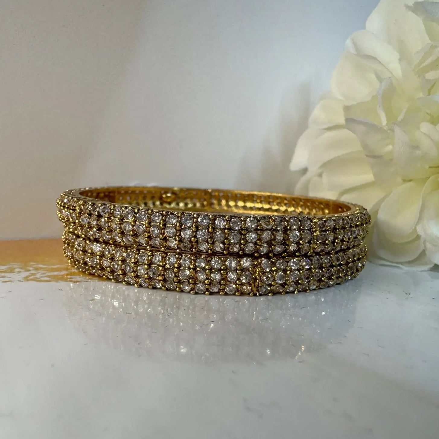 Gold plated bangles with clear gemstones on a white surface with flowers in the background