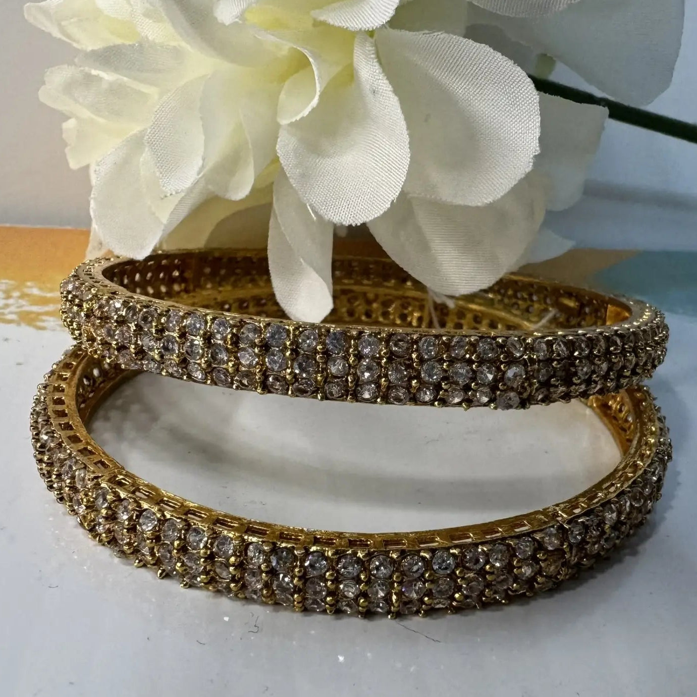 Gold plated bangles with clear gemstones and crystal embellishments on a white surface with a white flower in the background.