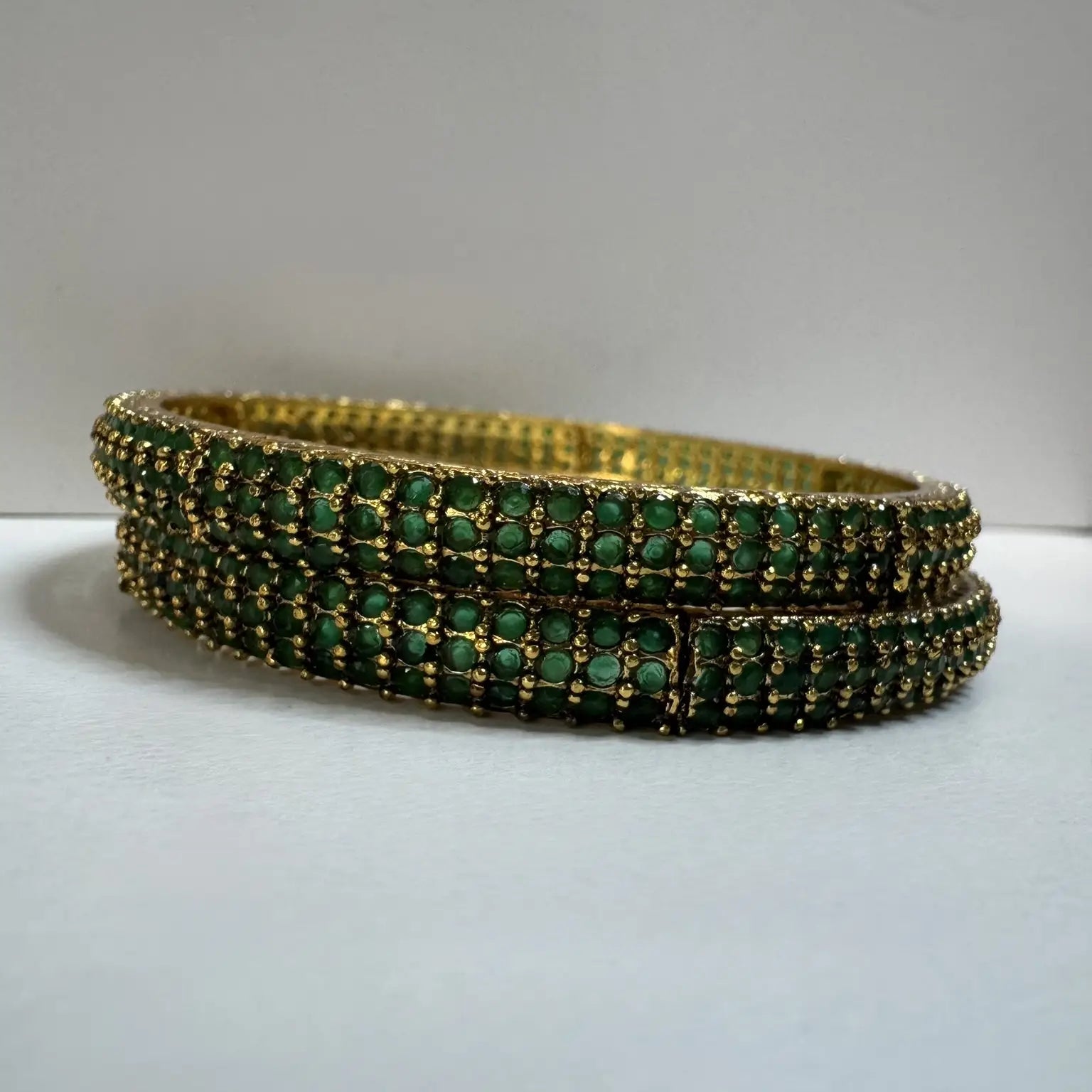 Gold plated bangles with green gemstones on a white surface with a gray background