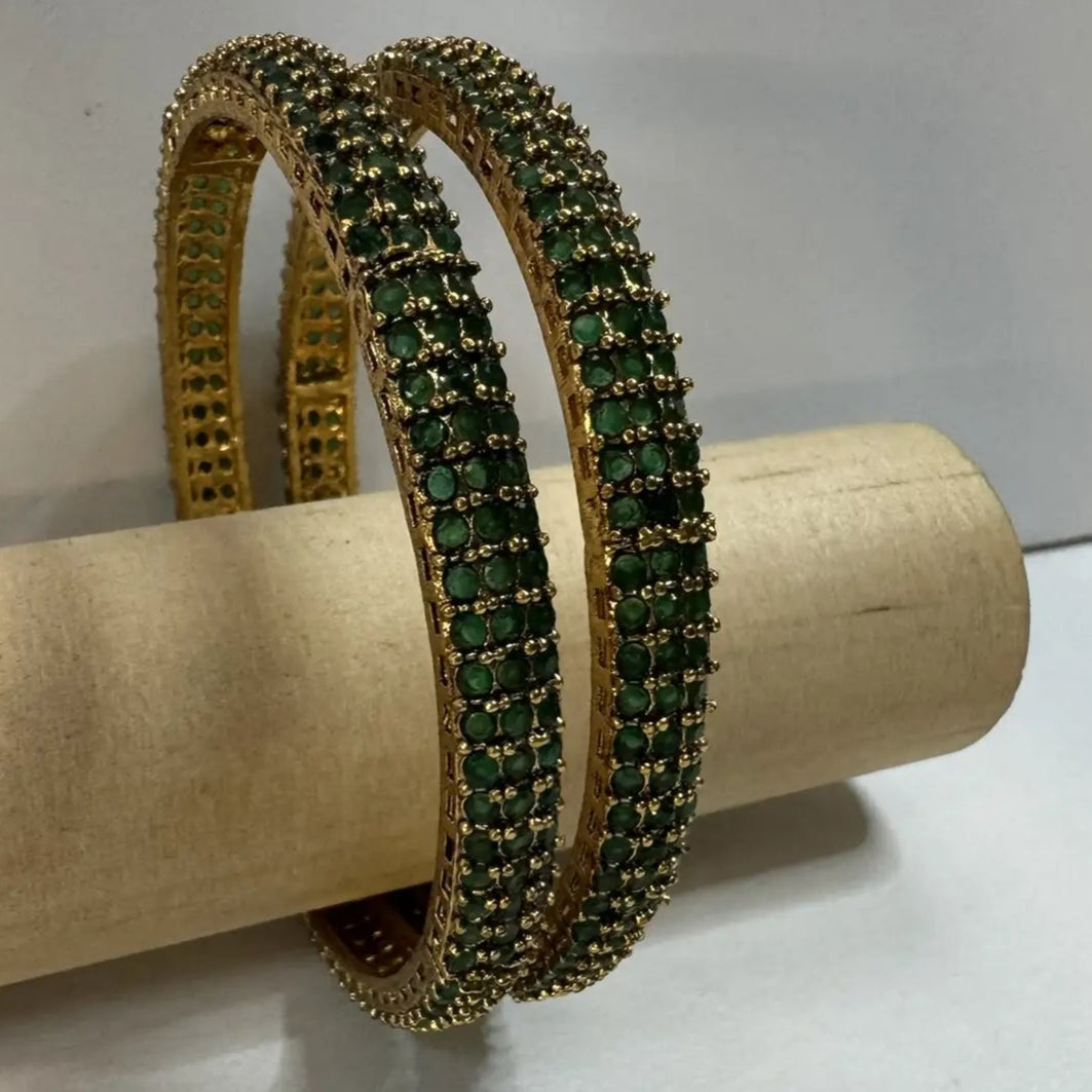 Gold plated bangles with green gemstones on a wooden roll against a white background