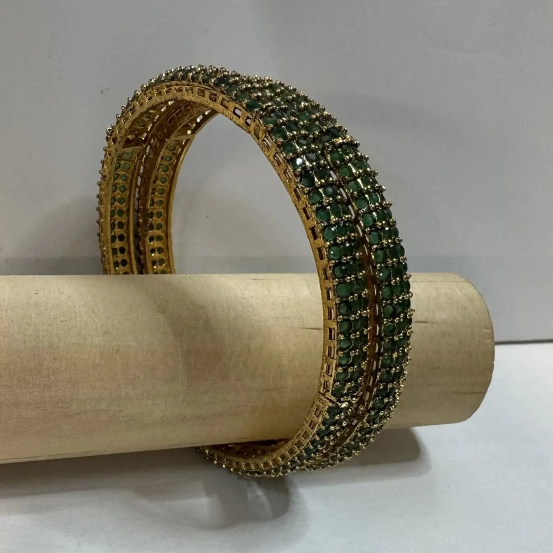 Gold plated bangles with green gemstones on a cylindrical wooden block against a white background