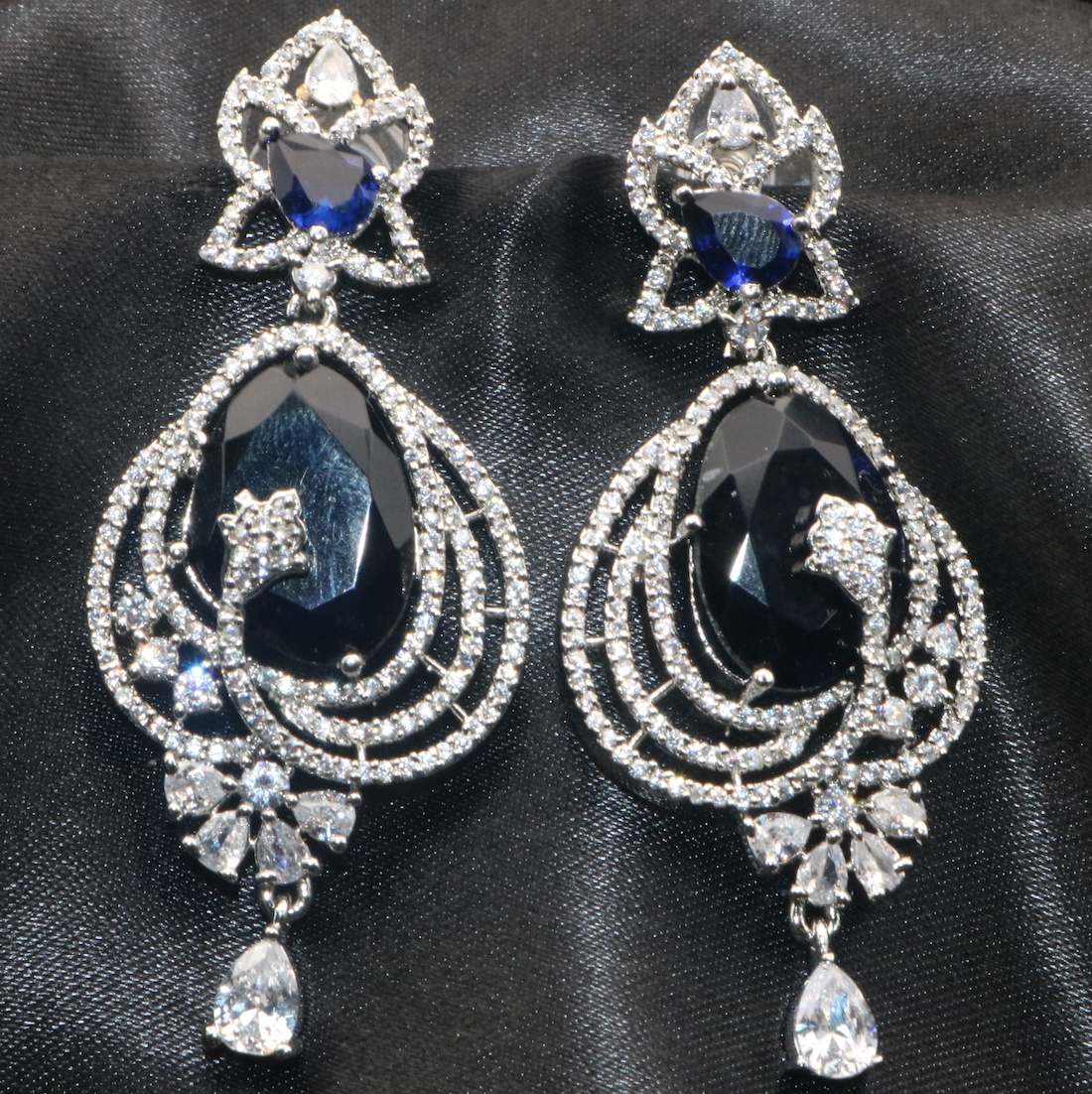 closer view of pair of stainless steel earrings with blue and clear embellishments, designed to resemble a flower or a geometric shape, set with American diamonds.