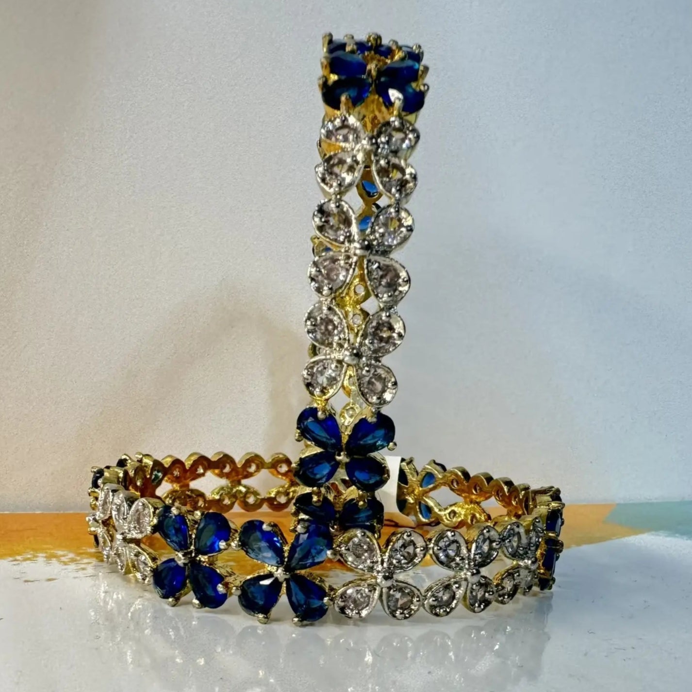 Gold plated bangles with blue and clear gemstones in a floral pattern on a light background