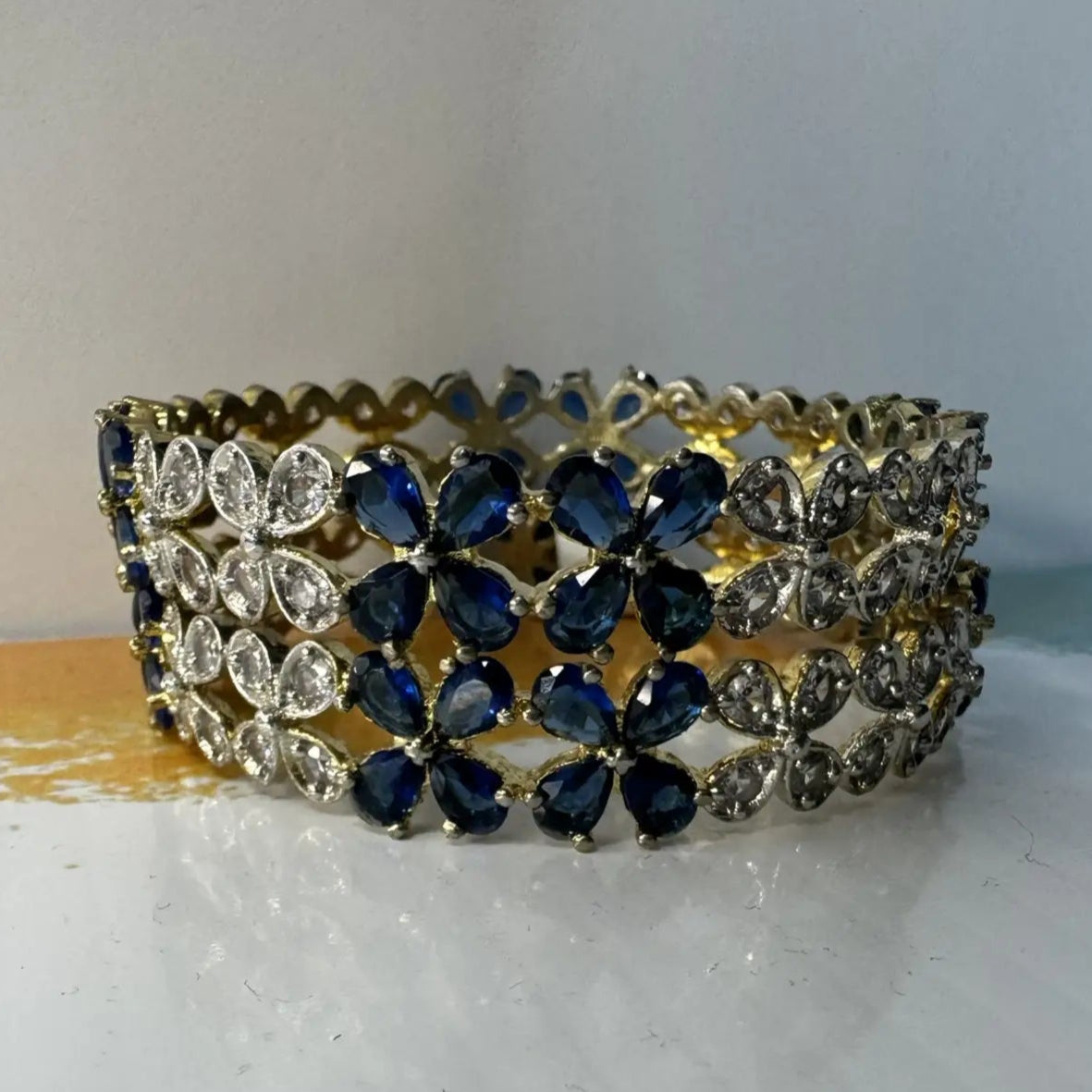 Gold plated bangles with blue and clear gemstones in a floral pattern and gold accents on a white surface