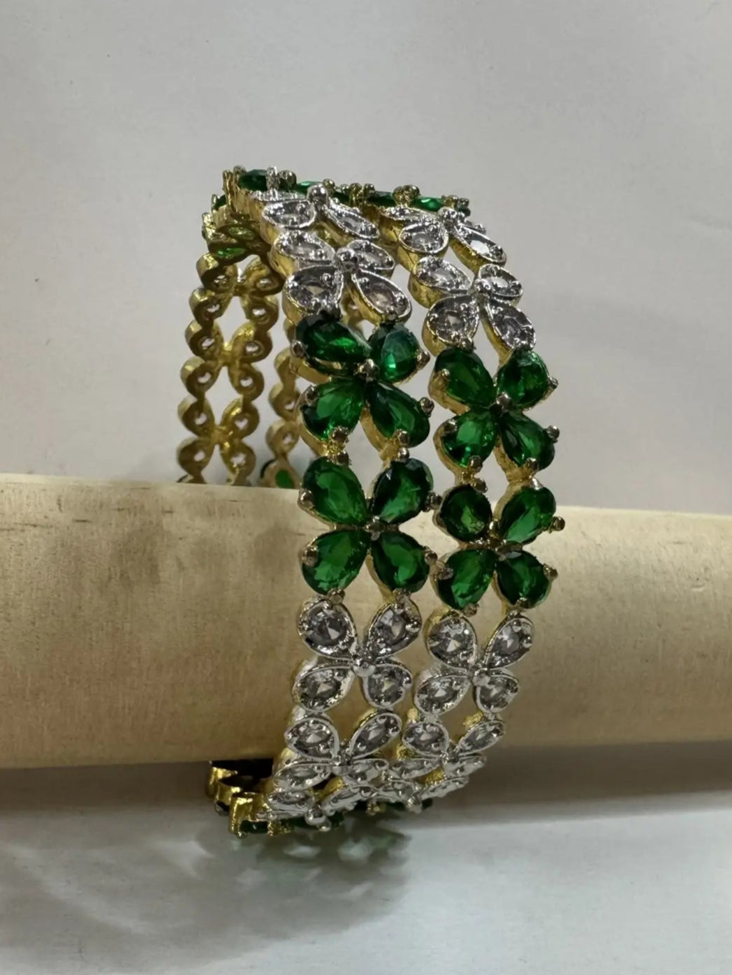 Gold plated bangles with green and clear gemstones in a floral pattern and gold accent on a wooden stick against a gray background