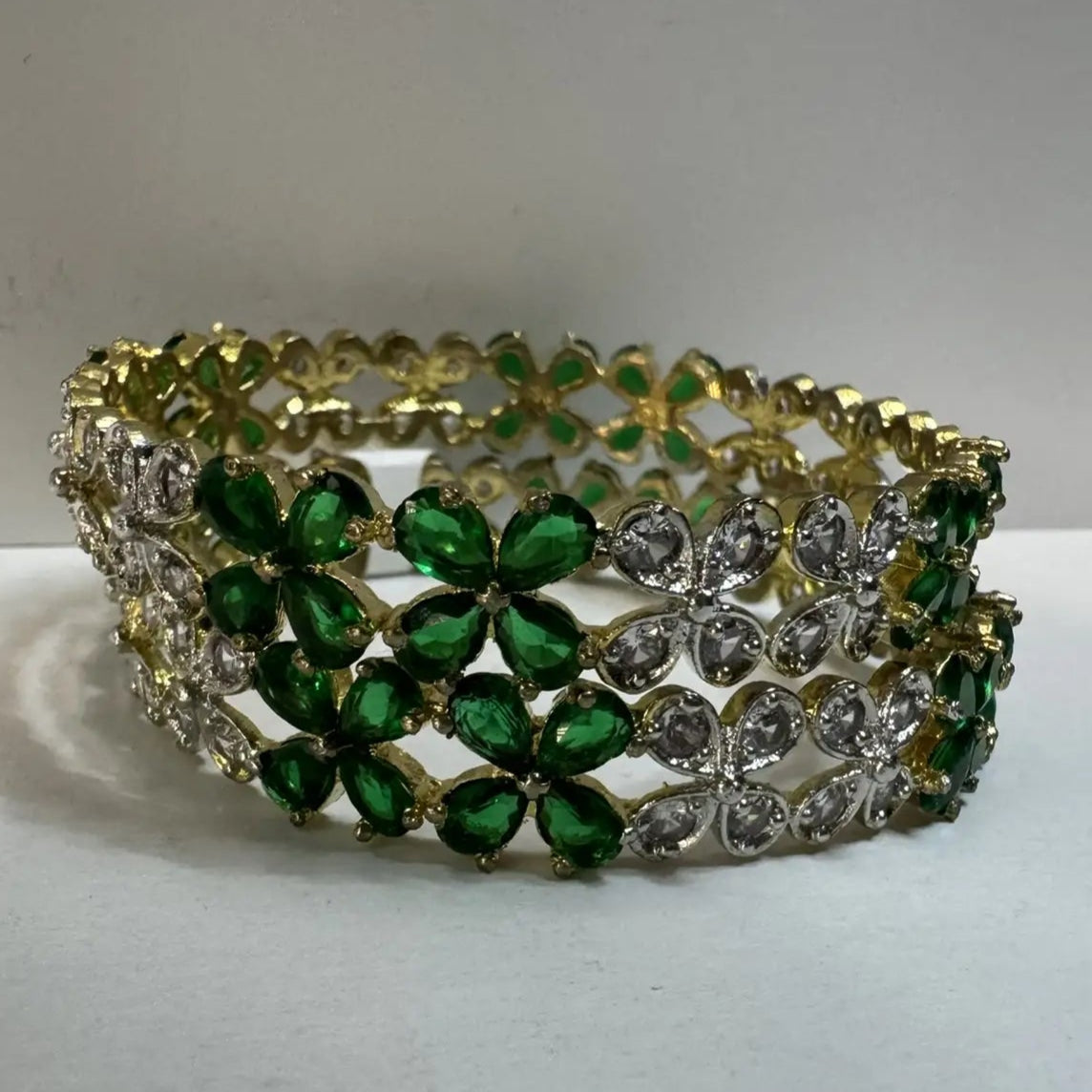 Gold plated bangles with green and clear gemstones in a floral pattern and gold accent on a white surface