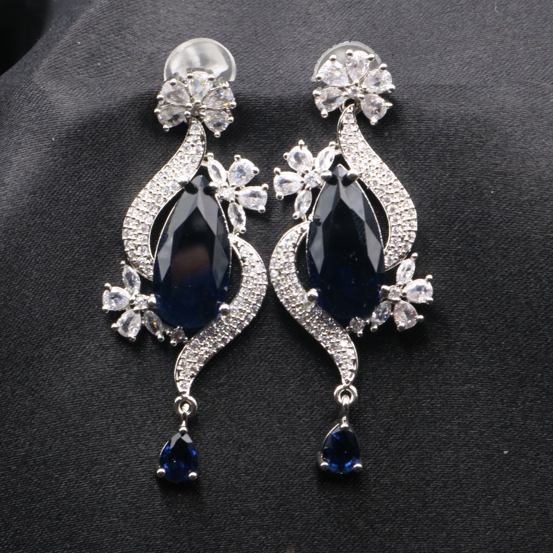 A pair of elegant ROYA earrings featuring stainless steel, American diamond accents, and blue gemstones, displayed against a dark background.