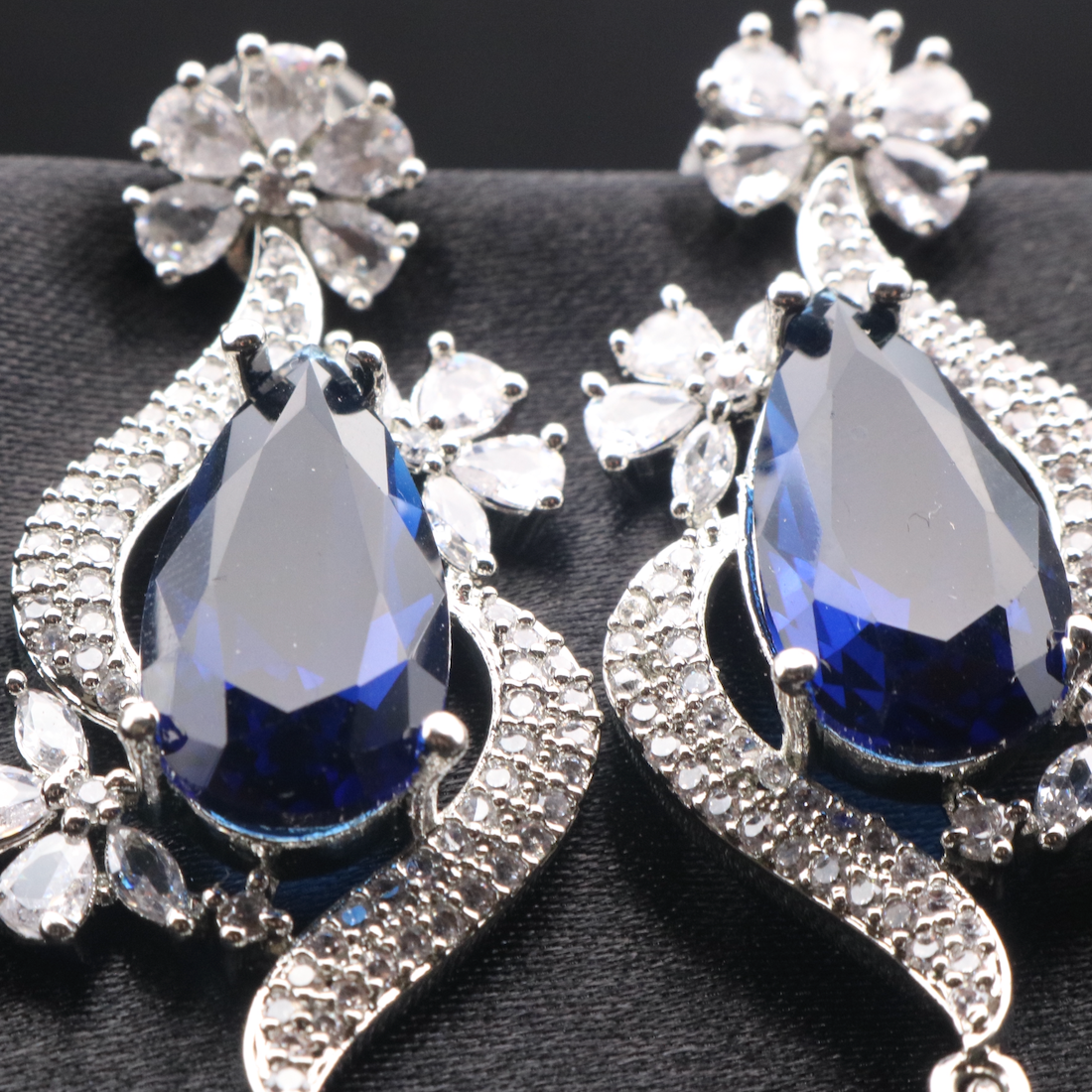 closer view of pair of elegant ROYA earrings featuring stainless steel, American diamond accents, and blue gemstones, displayed against a dark background.