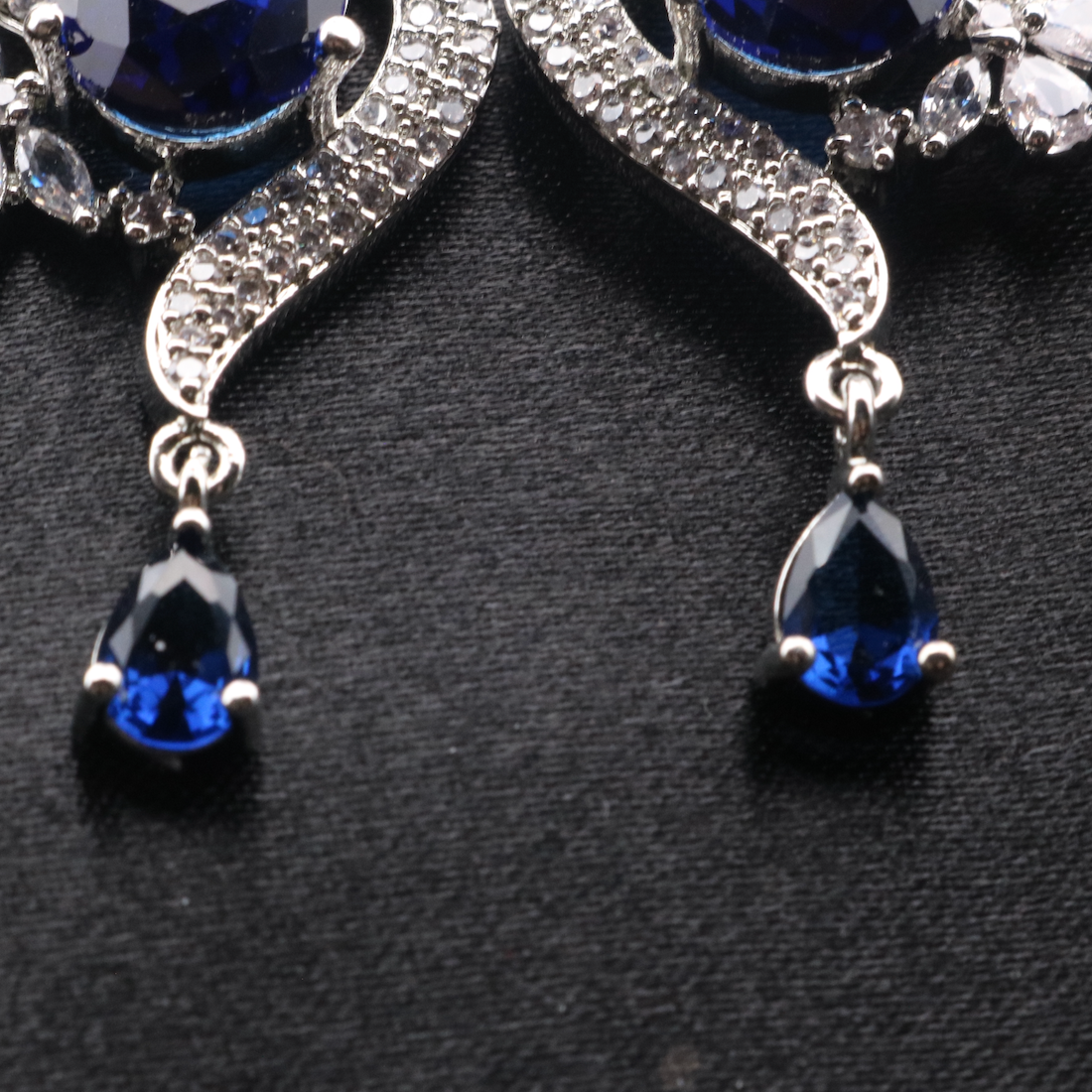 bottom view of pair of elegant ROYA earrings featuring stainless steel, American diamond accents, and blue gemstones, displayed against a dark background.