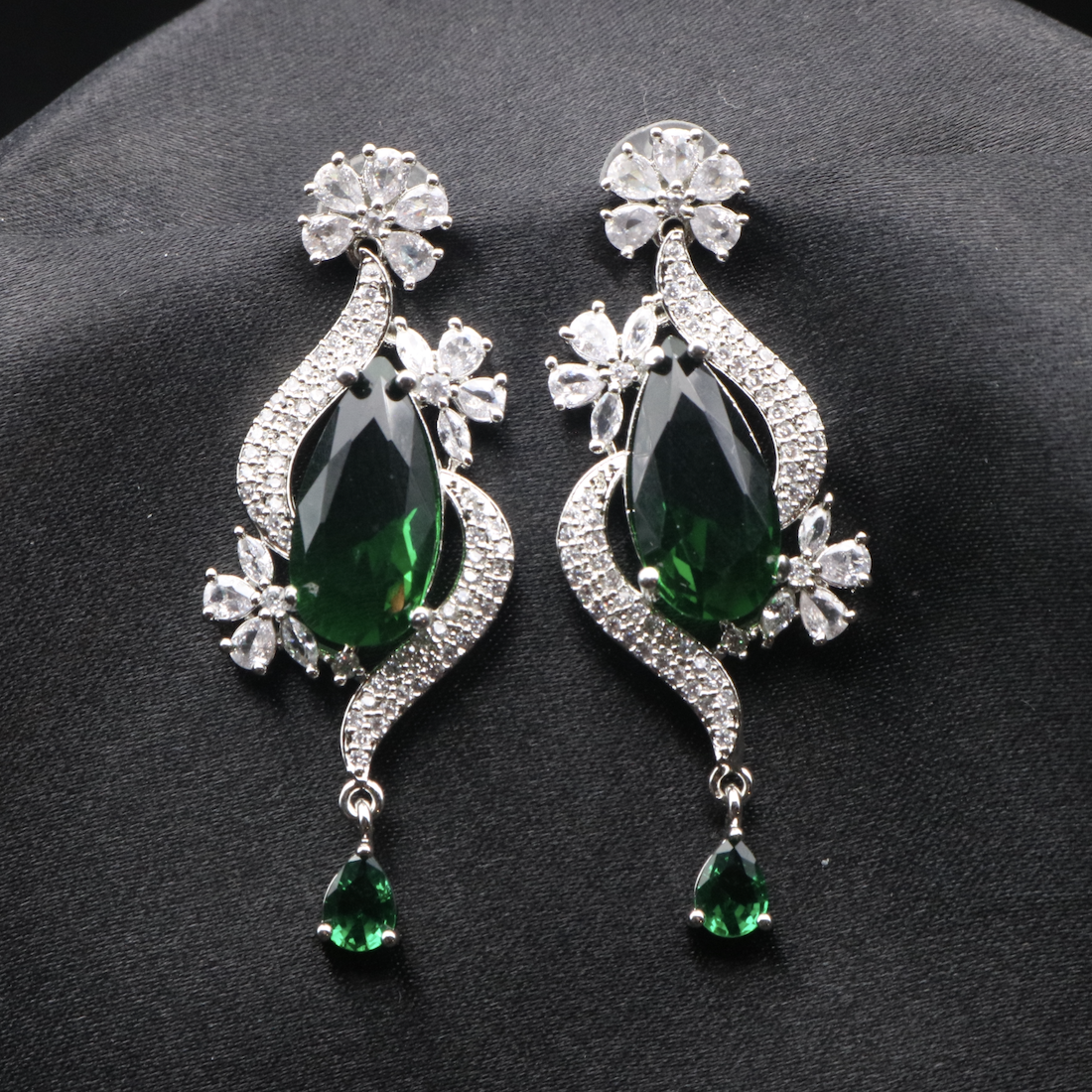 A pair of elegant ROYA earrings featuring stainless steel, American diamond accents, and green gemstones, displayed against a dark background.