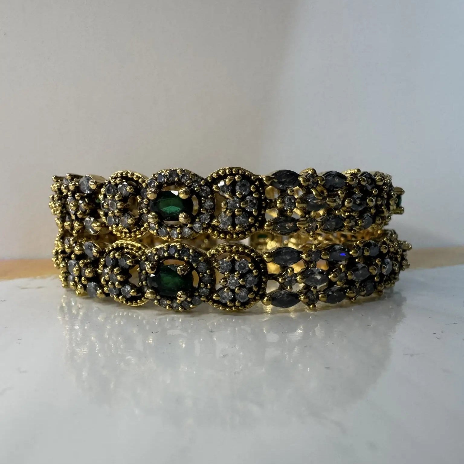 A set of gold-plated stainless steel bangles adorned with American diamond accents and green gemstones on a white surface