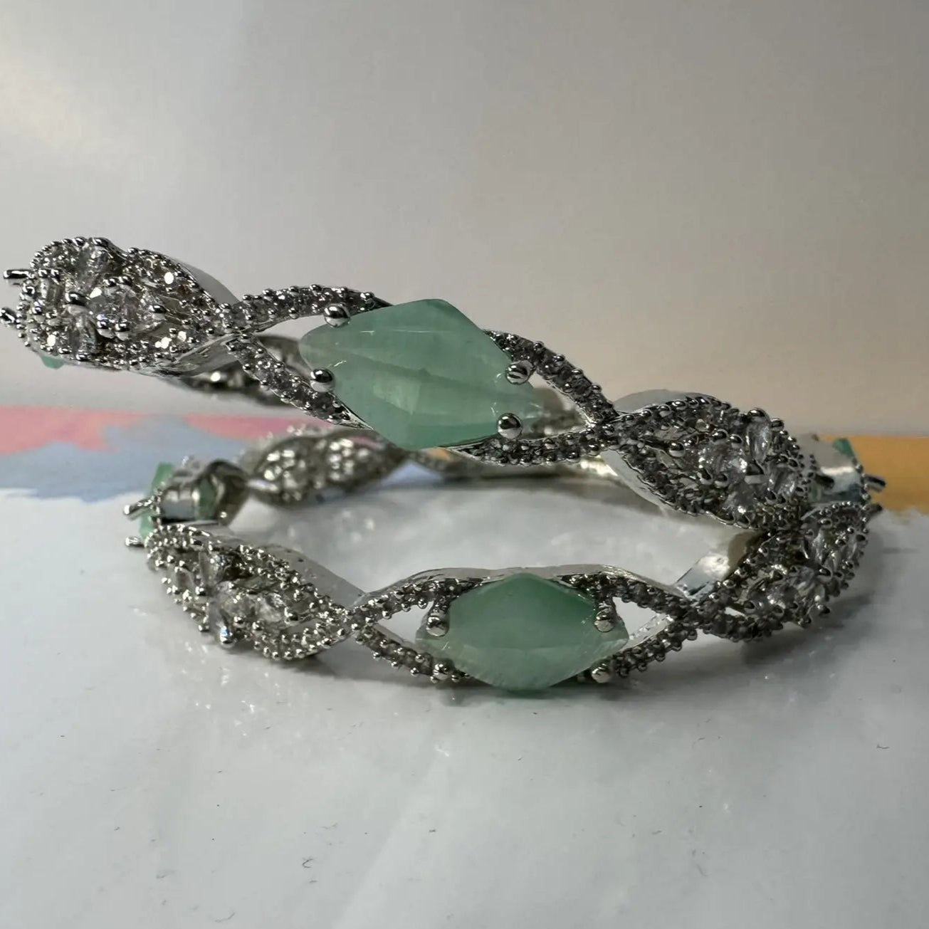 A pair of silver bangles adorned with turquoise rhinestones and American Diamond encrusted on a silver base, displayed on a white surface and white background