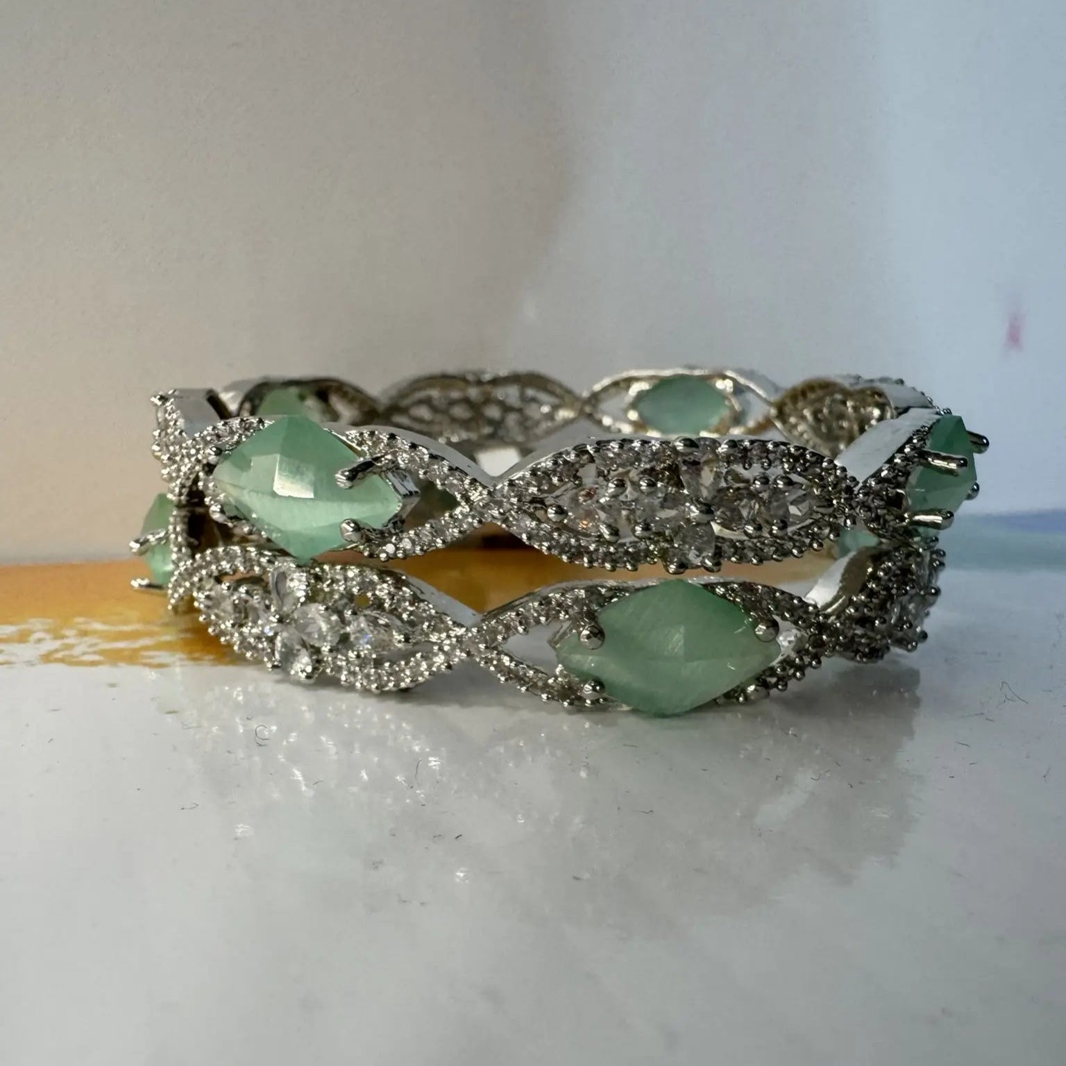 A pair of silver bangles adorned with turquoise rhinestones and American Diamond encrusted on a silver base, displayed on a white surface.