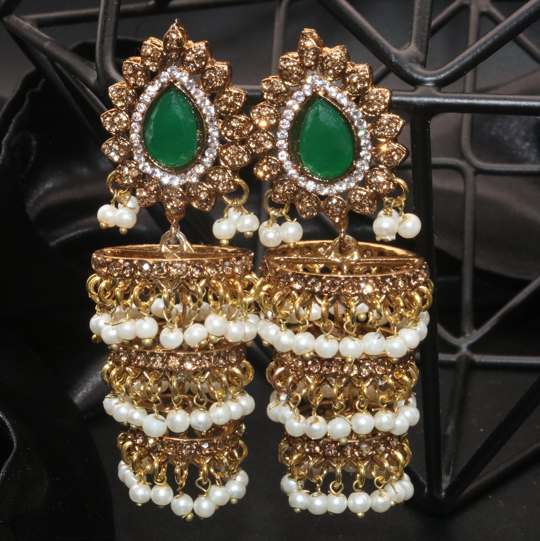 A pair of gold-colored jhumka earrings with white pearl-like beads and green gemstone accents, displayed against a black background.